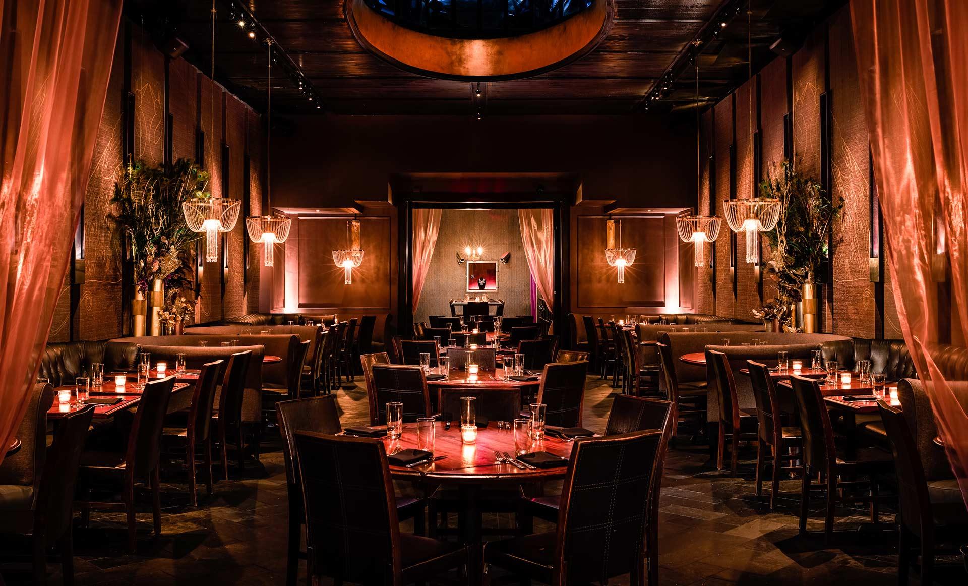 A dimly lit, elegant restaurant with dark wood tables, cushioned chairs, warm pendant lighting, and soft drapes, creating an intimate and upscale atmosphere.