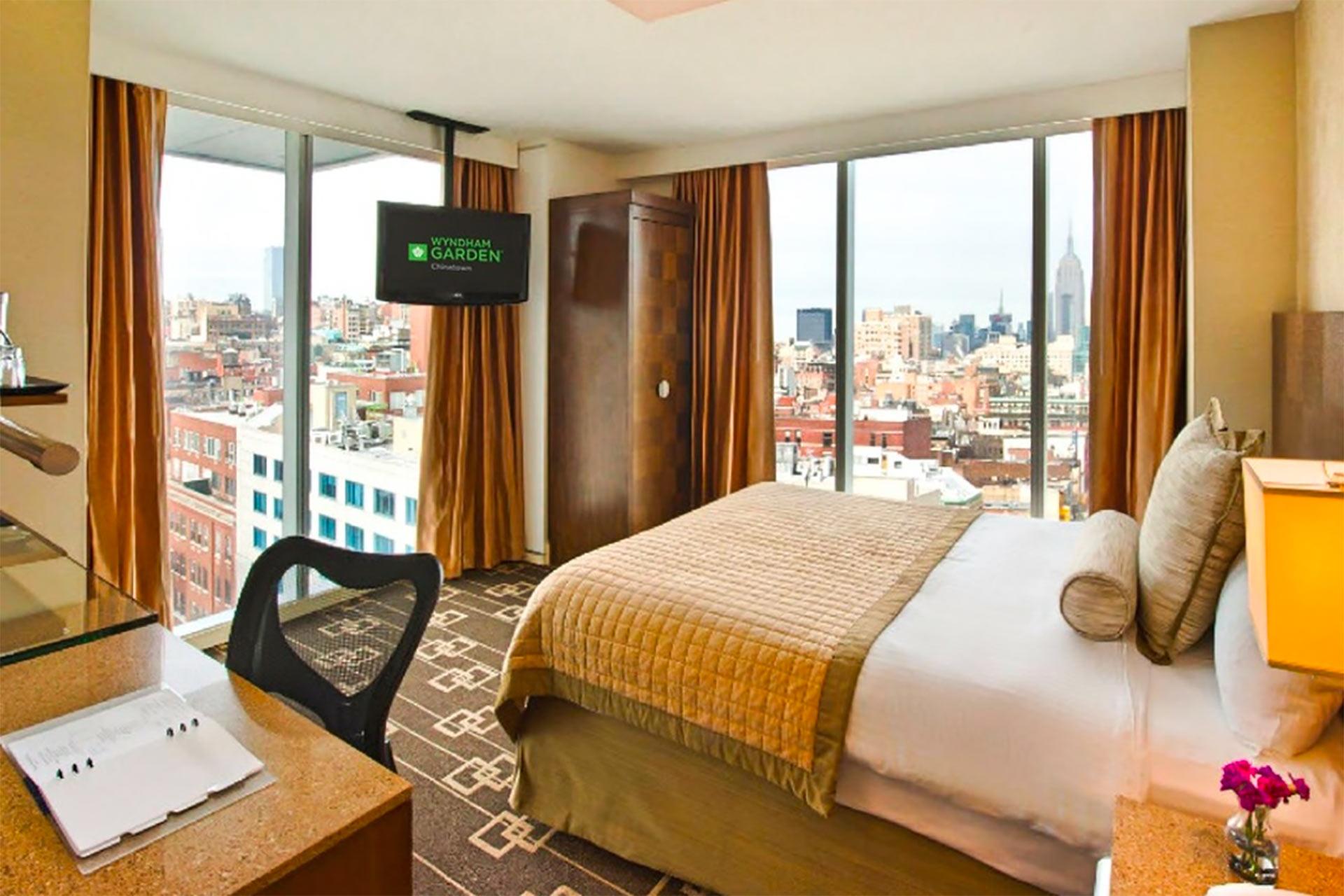 A modern hotel room with a large bed, desk, chair, TV, and floor-to-ceiling windows showing a city skyline with tall buildings and a cloudy sky. Gold curtains frame the windows, and a small flower vase sits on the nightstand.