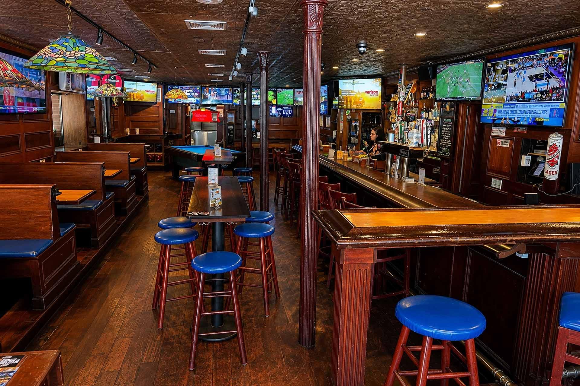 A sports bar interior with wooden booths, blue cushioned stools, several tables, multiple TVs displaying sports, a bar counter with drinks, and a stained-glass ceiling lamp; the space appears empty of customers.