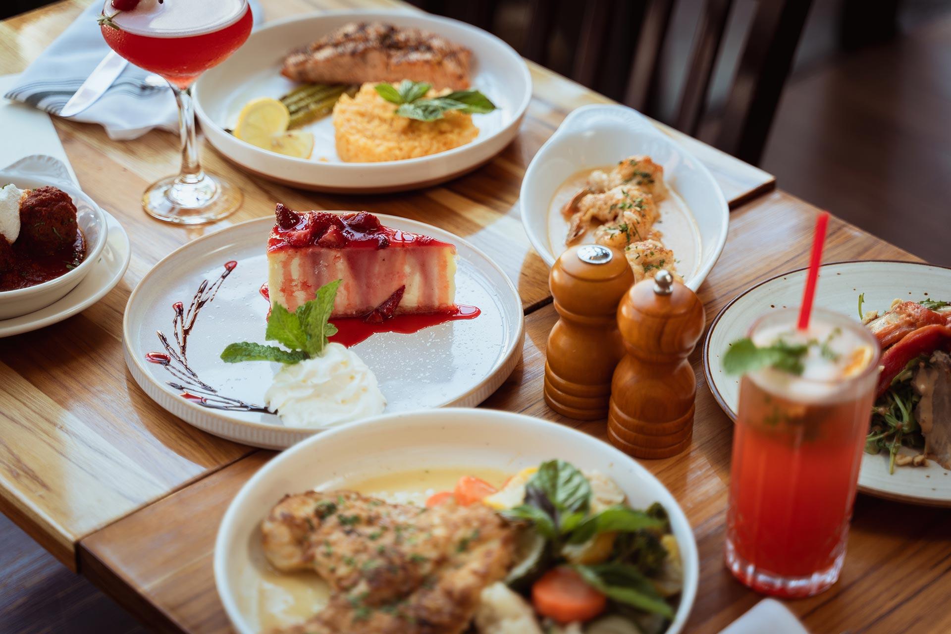 A wooden table set with plates of various meals, a slice of cheesecake with berry sauce and whipped cream, a red cocktail, a pink drink with a straw, and salt and pepper shakers.