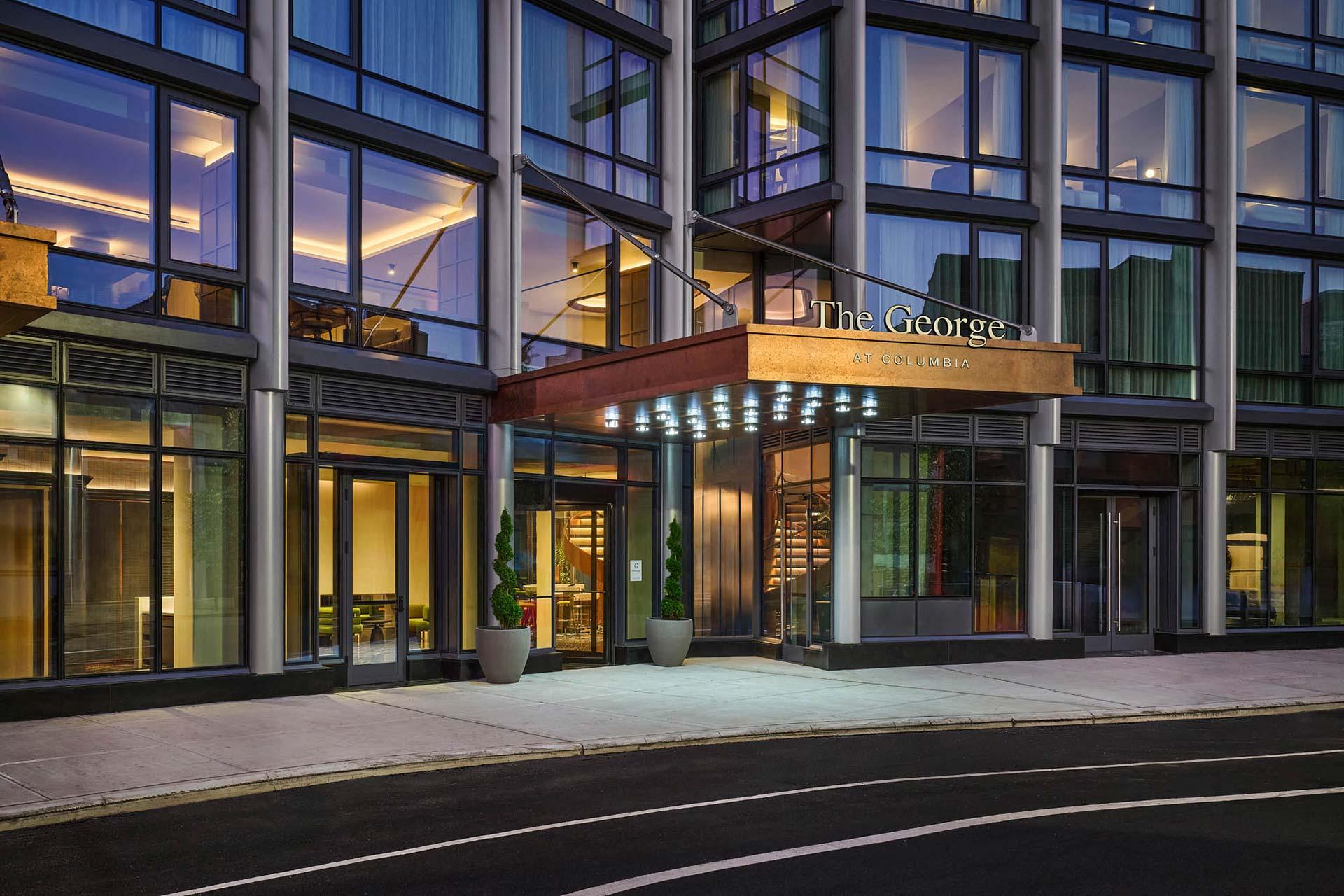 The exterior of The George, a modern building with large glass windows, illuminated entrance, and potted plants by the doorway, taken at dusk.