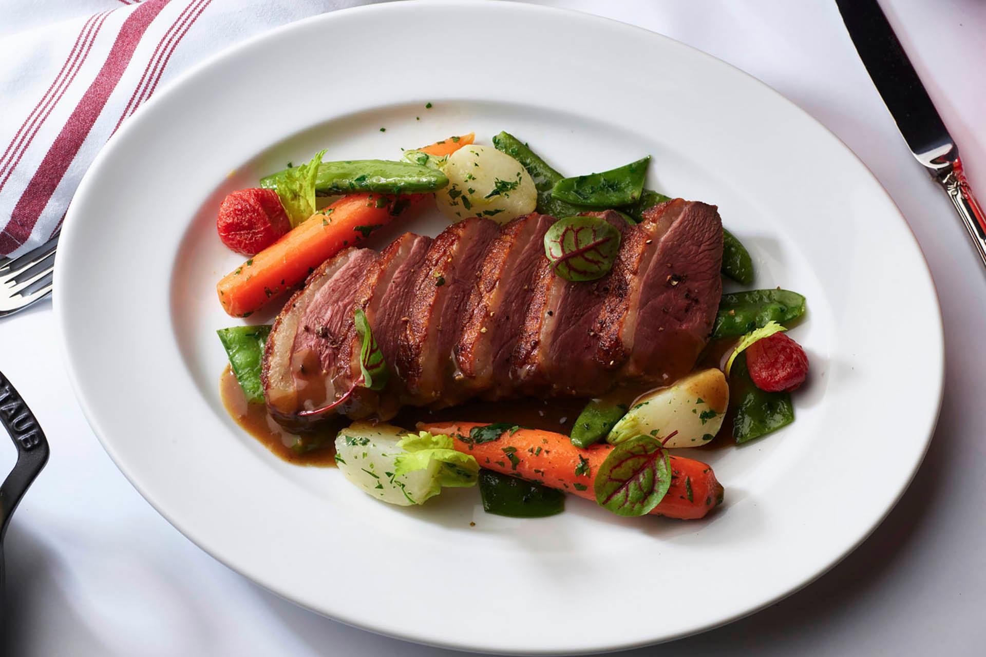 A white plate with sliced roast beef, glazed carrots, baby potatoes, sugar snap peas, and cherry tomatoes, garnished with herbs, resting on a white tablecloth with a fork and knife beside it.