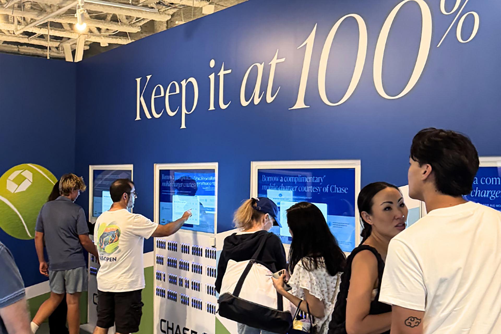 People stand in line at a wall with the words "Keep it at 100%" above several digital screens, possibly at a promotional or informational booth, with a tennis-themed design and the Chase logo visible.