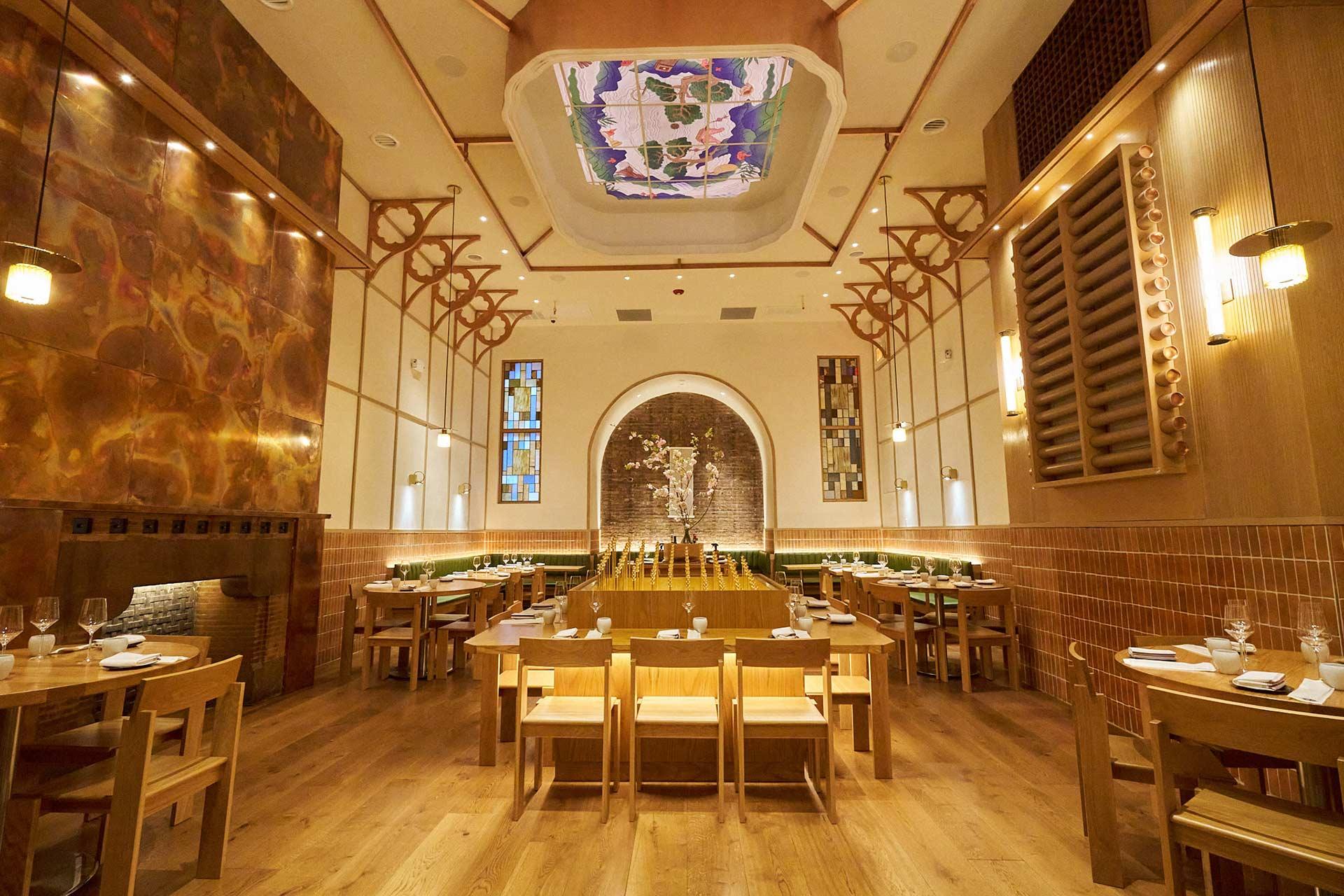A modern restaurant interior with wooden tables and chairs, warm lighting, and an artistic ceiling mural. The room features a central arched wall with a textured art display and a spacious, inviting atmosphere.