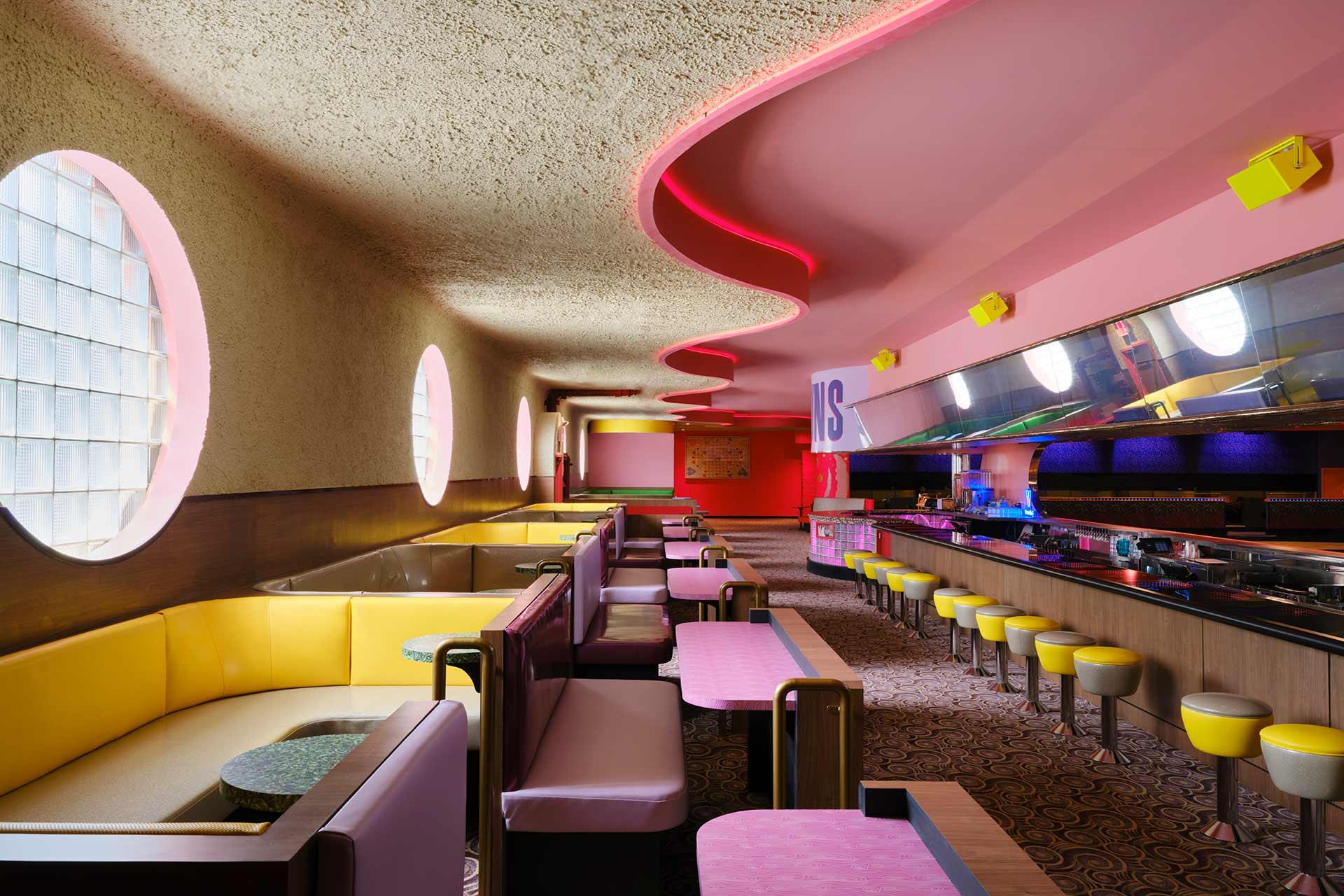 Retro-style diner with curved pink ceiling lights, yellow stools at a counter, pink tables, circular windows, and booths with yellow and brown seating. The decor features bold colors and modern, playful design elements.