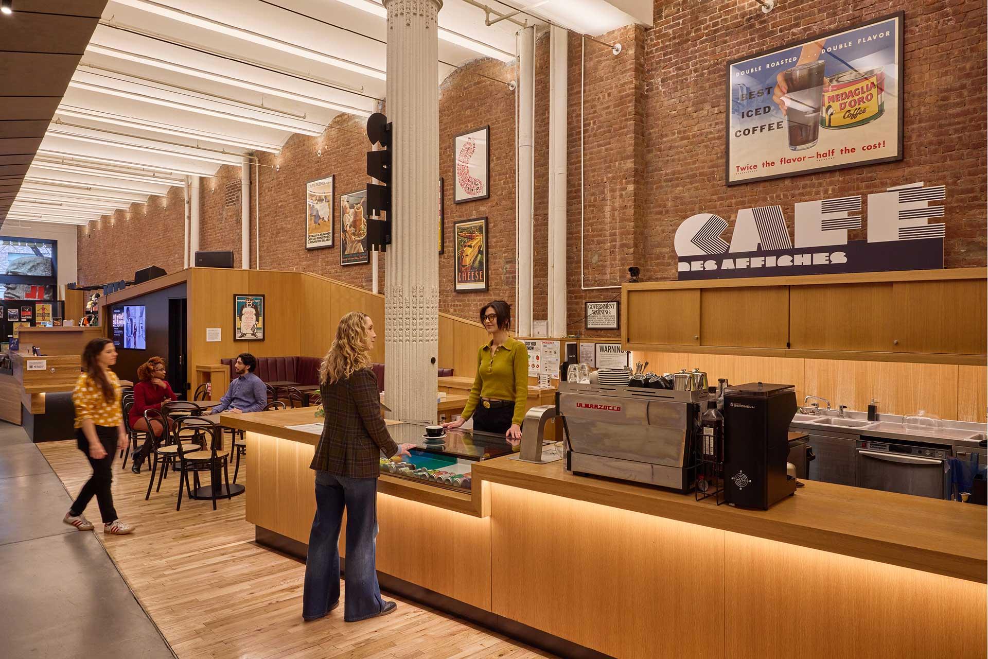 People interact at the counter of a modern café with brick walls, large posters, and high ceilings. The space features wooden accents, a coffee machine, and customers seated at tables.