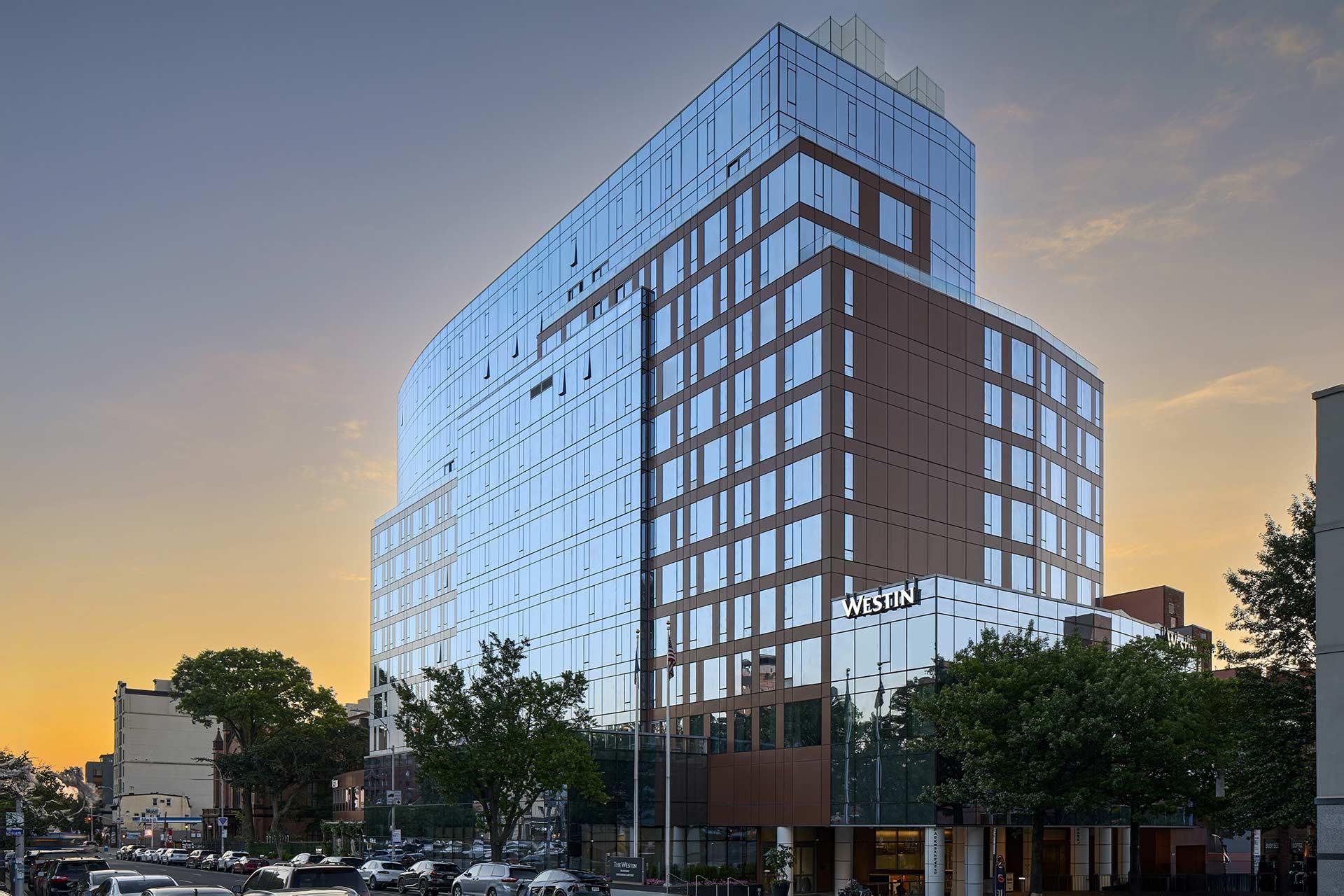 A modern, glass-fronted high-rise hotel building with "Westin" signage, set against a sunset sky. Trees and cars line the busy street in front of the hotel.