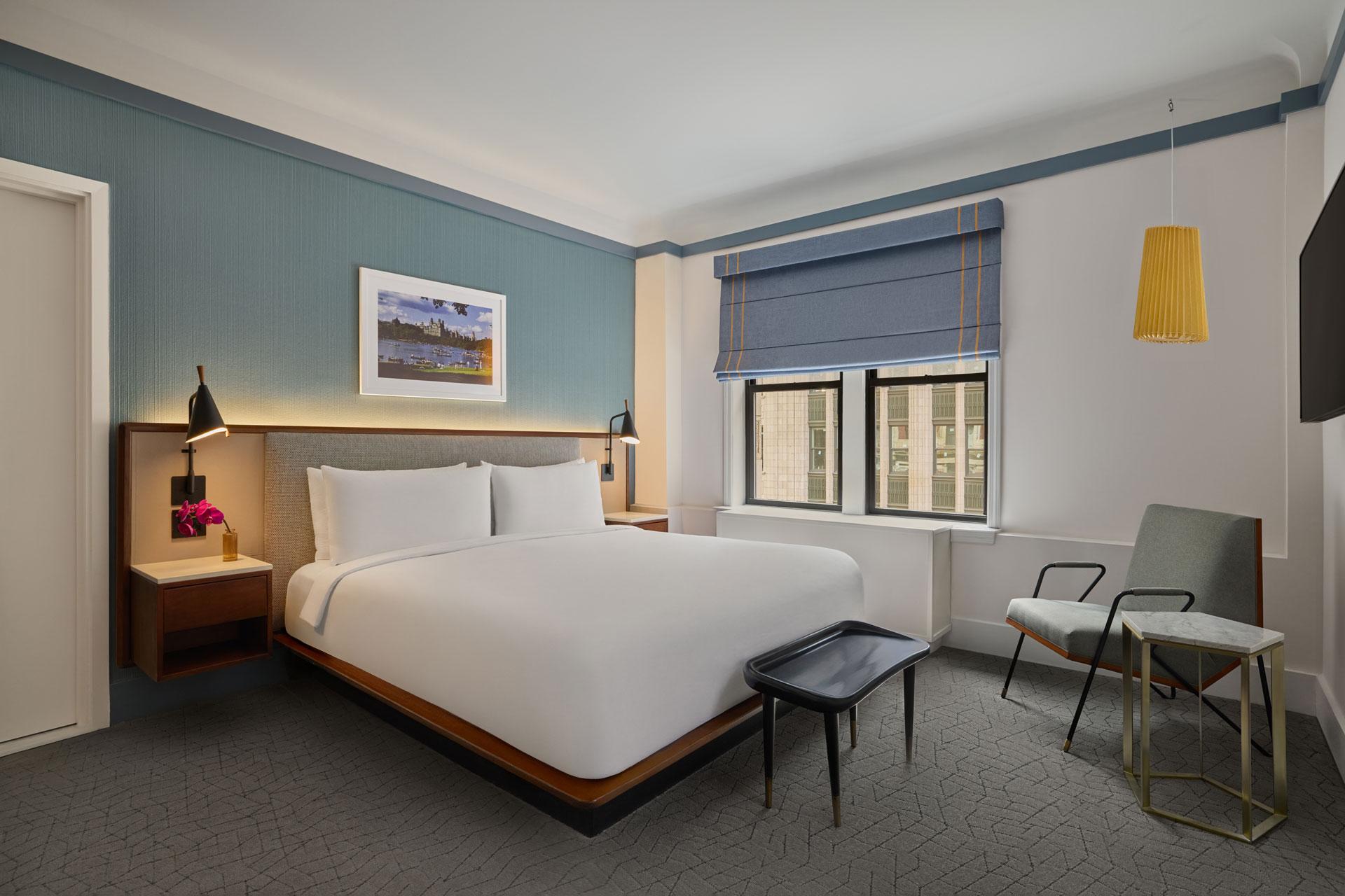 Modern hotel room with a king bed, two nightstands, lamps, wall art, a window with blue blinds, a chair, a small table, and a bench at the foot of the bed. The decor is minimalist and inviting.