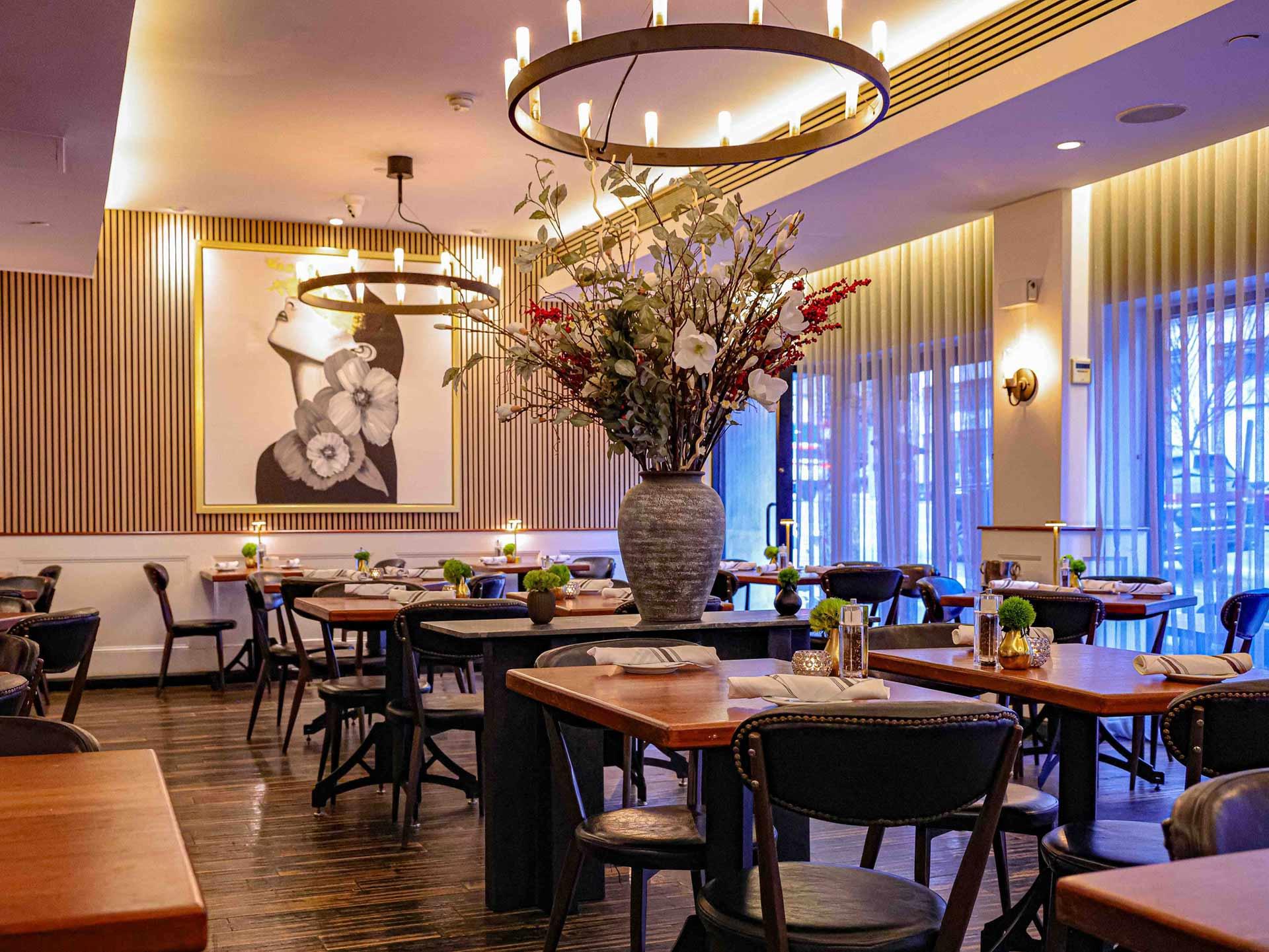 A stylish, modern restaurant interior with wooden tables, black chairs, a large floral arrangement in the center, soft lighting, and a portrait of a woman on the wall. Floor-to-ceiling windows let in natural light.