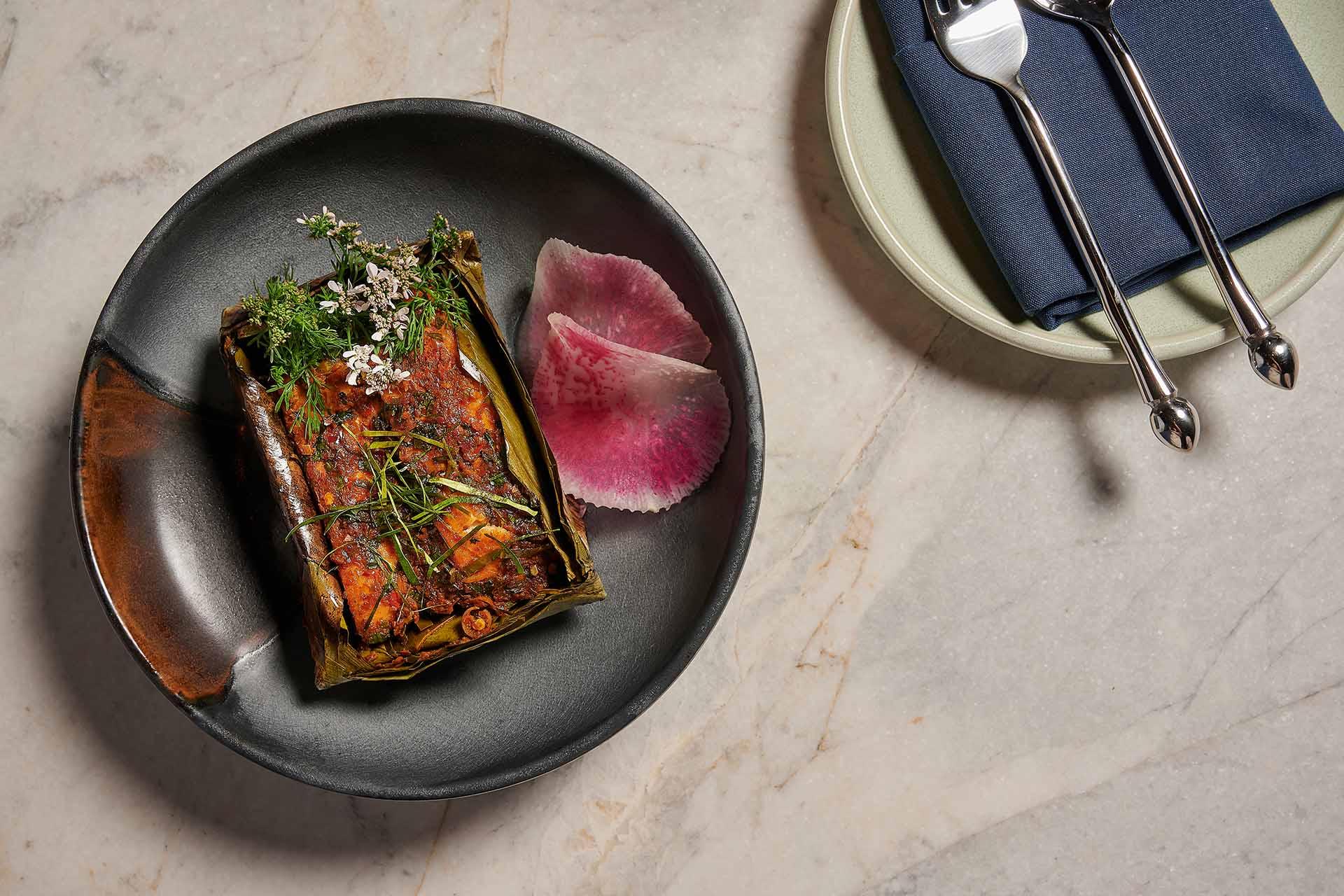 A black plate with grilled food wrapped in leaves, garnished with herbs and flowers, next to two thin slices of pink radish. Fork and spoon on a navy napkin are placed on a small dish nearby.