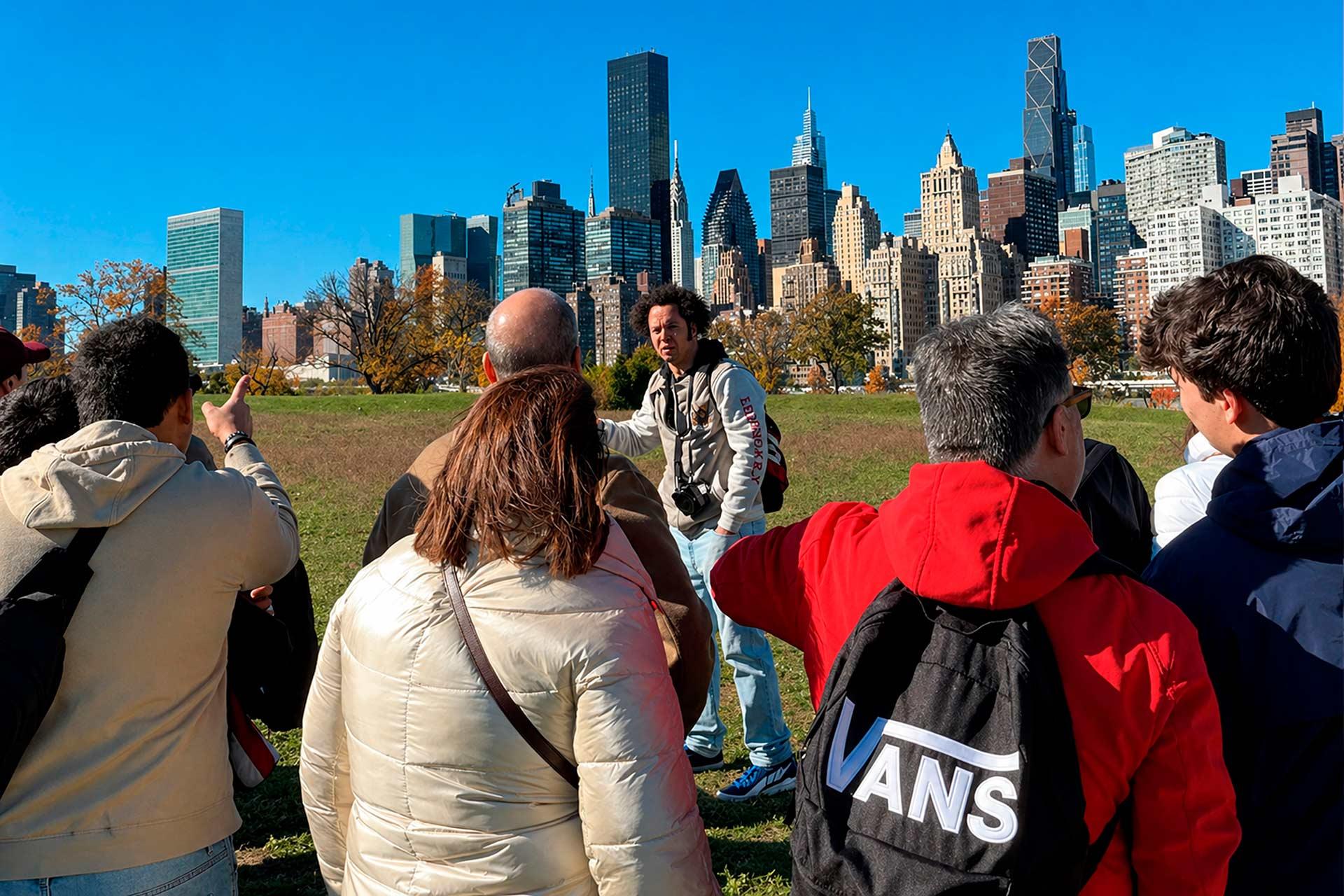 A group of people on a grassy area listens to a tour guide with New York City skyscrapers in the background under a clear blue sky. One person has a black backpack with a visible "VANS" logo.