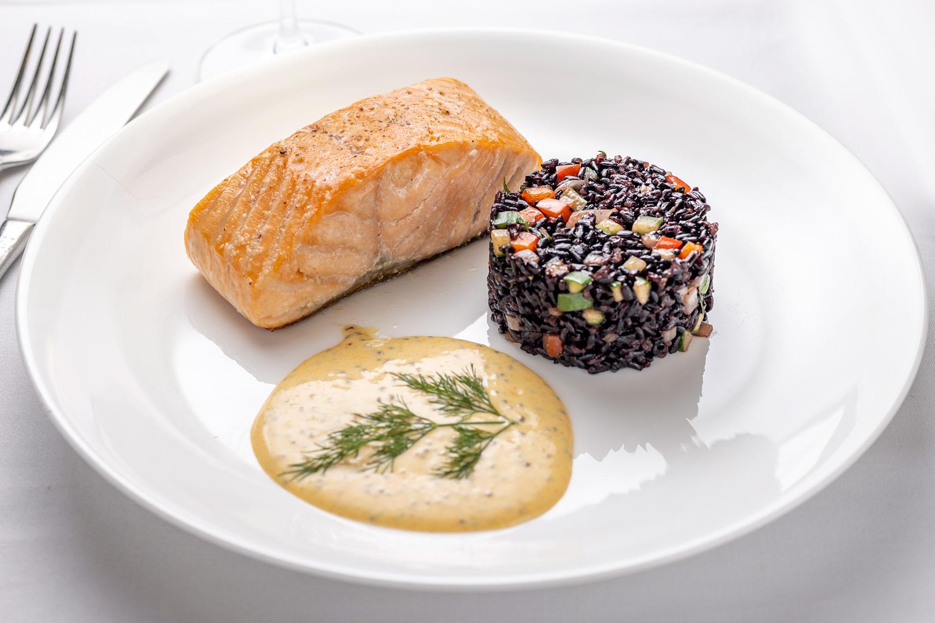 A plate with a piece of cooked salmon, a round serving of black rice mixed with diced vegetables, and a portion of creamy yellow sauce garnished with dill, all arranged neatly on a white dish.