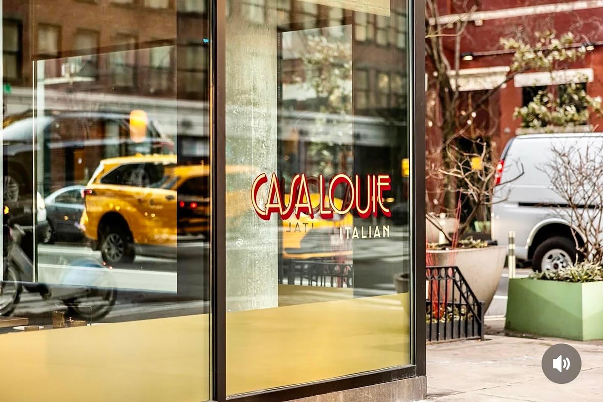 A glass window of an Italian restaurant named CasaLouie, reflecting a yellow taxi, cars, and city buildings on a sunny urban street.