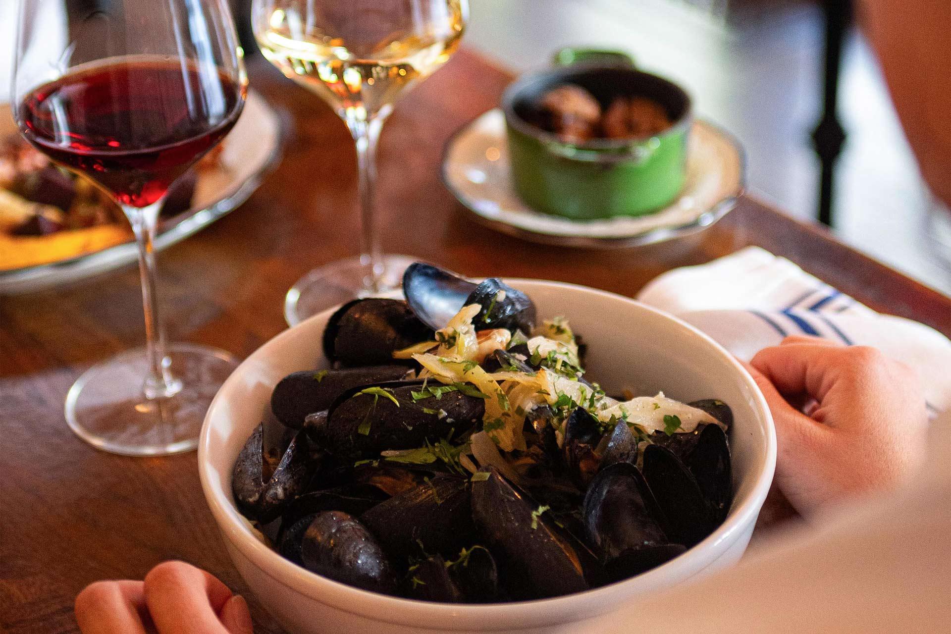 A person sits at a wooden table with a bowl of mussels garnished with herbs and onions, flanked by glasses of red and white wine and a small green pot of food in the background.