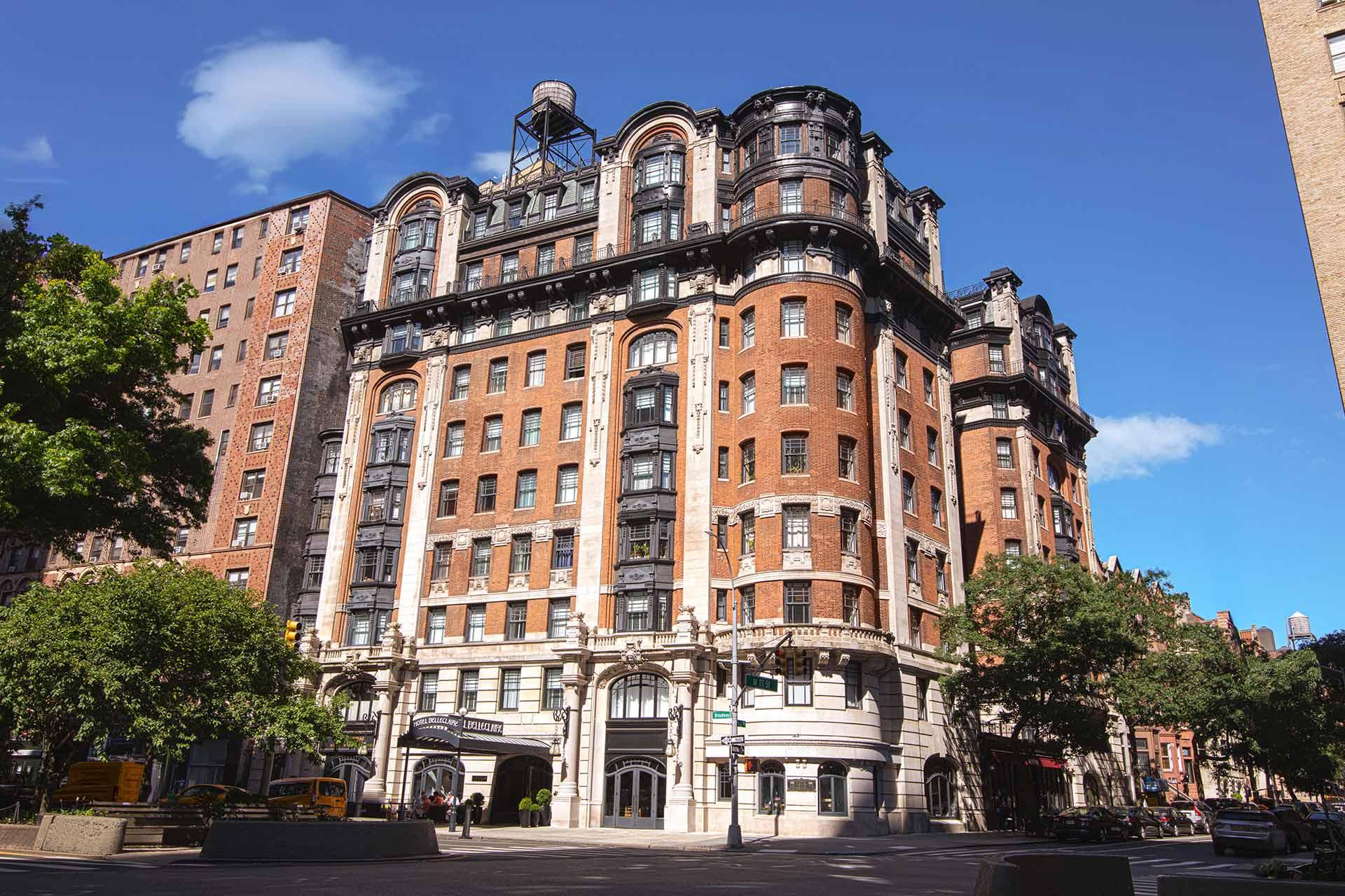 A historic, multi-story brick building with ornate architectural details stands on a sunny street corner, surrounded by trees and other buildings under a bright blue sky with a few clouds.