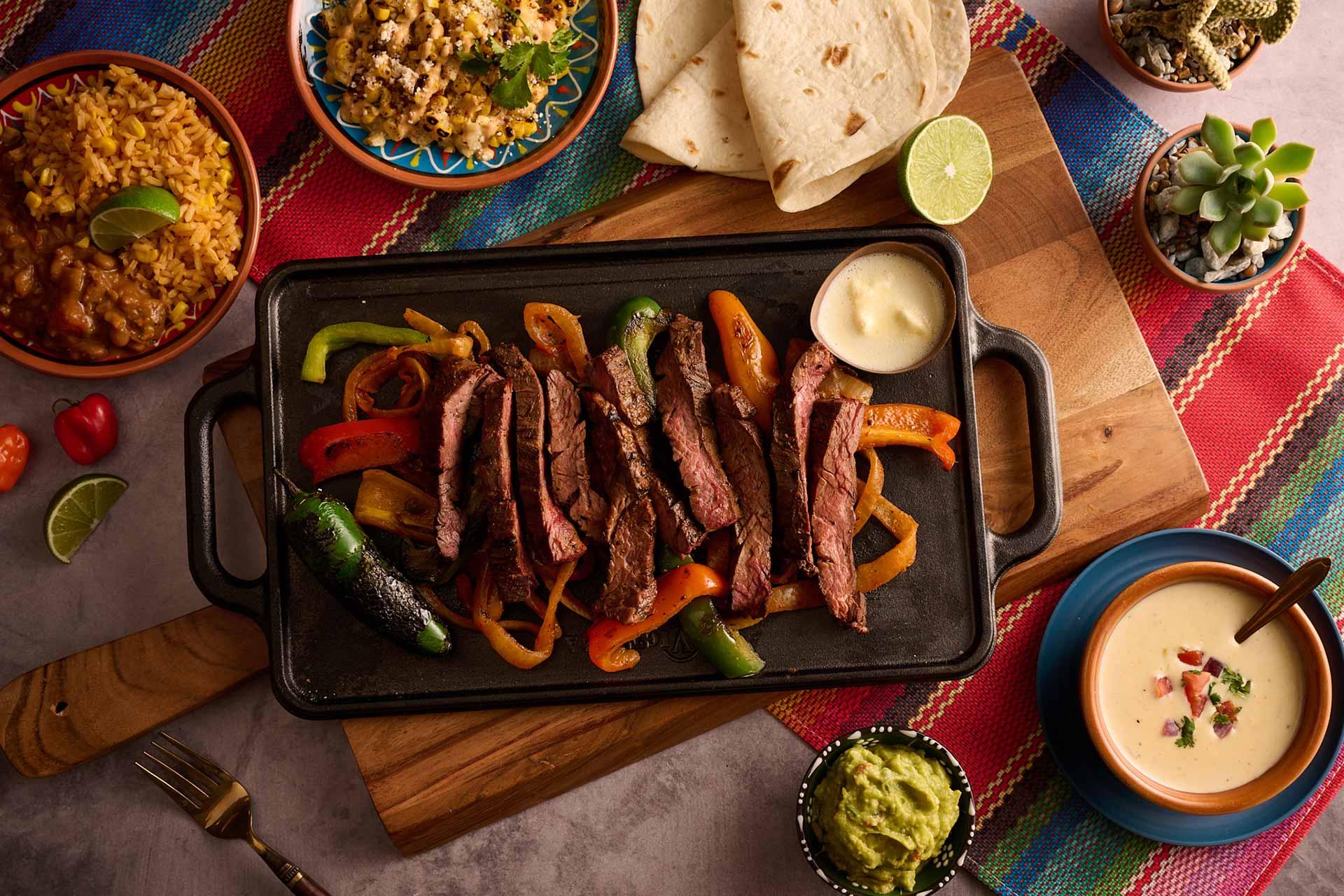 A sizzling beef fajita platter with grilled peppers and onions, surrounded by tortillas, rice, beans, queso dip, guacamole, and toppings on a colorful tablecloth.