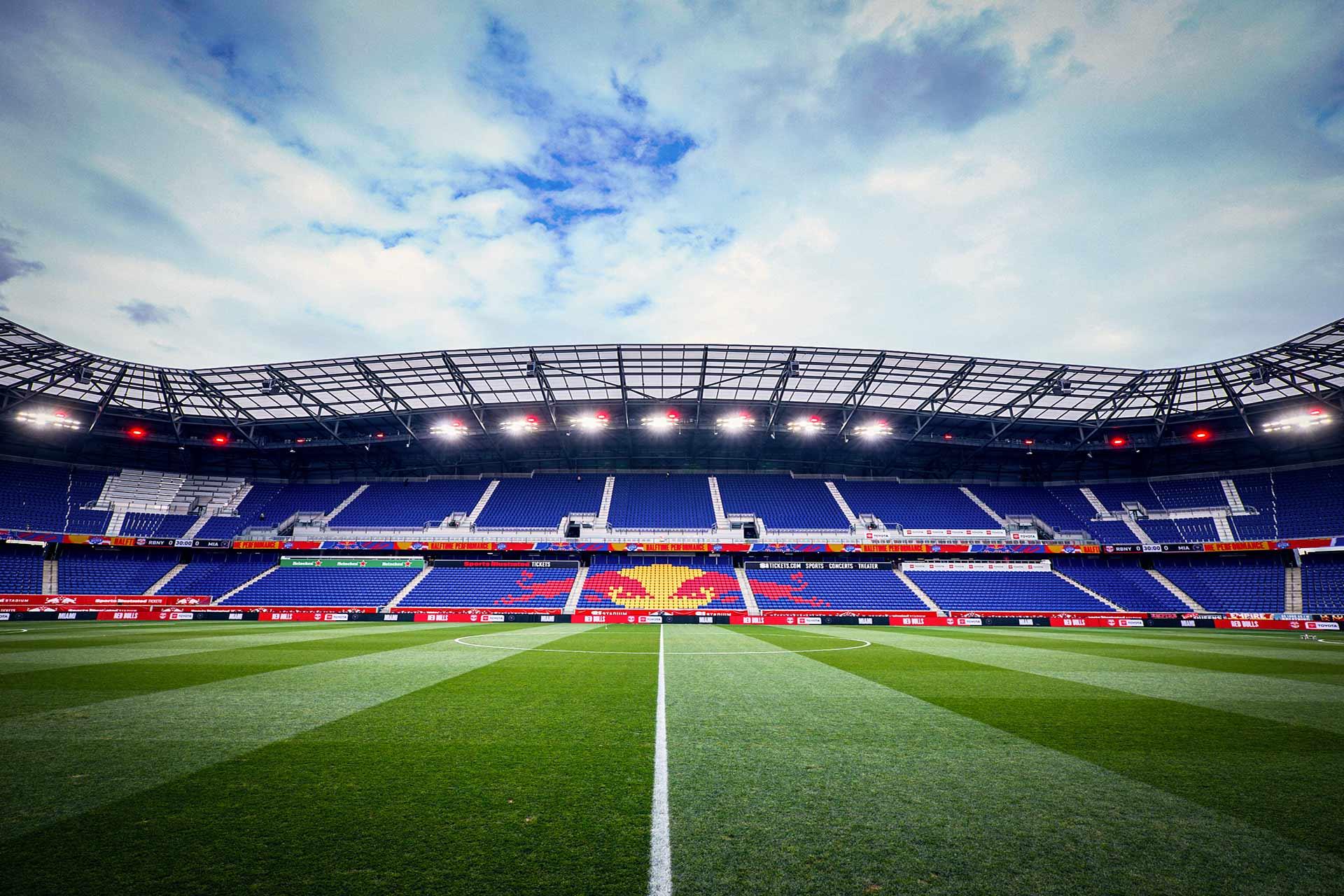 A view of an empty soccer stadium with blue seats, a large lion emblem at the center, and a bright green field under a partly cloudy sky. The stadium lights are on, and the perspective is from the center of the field.