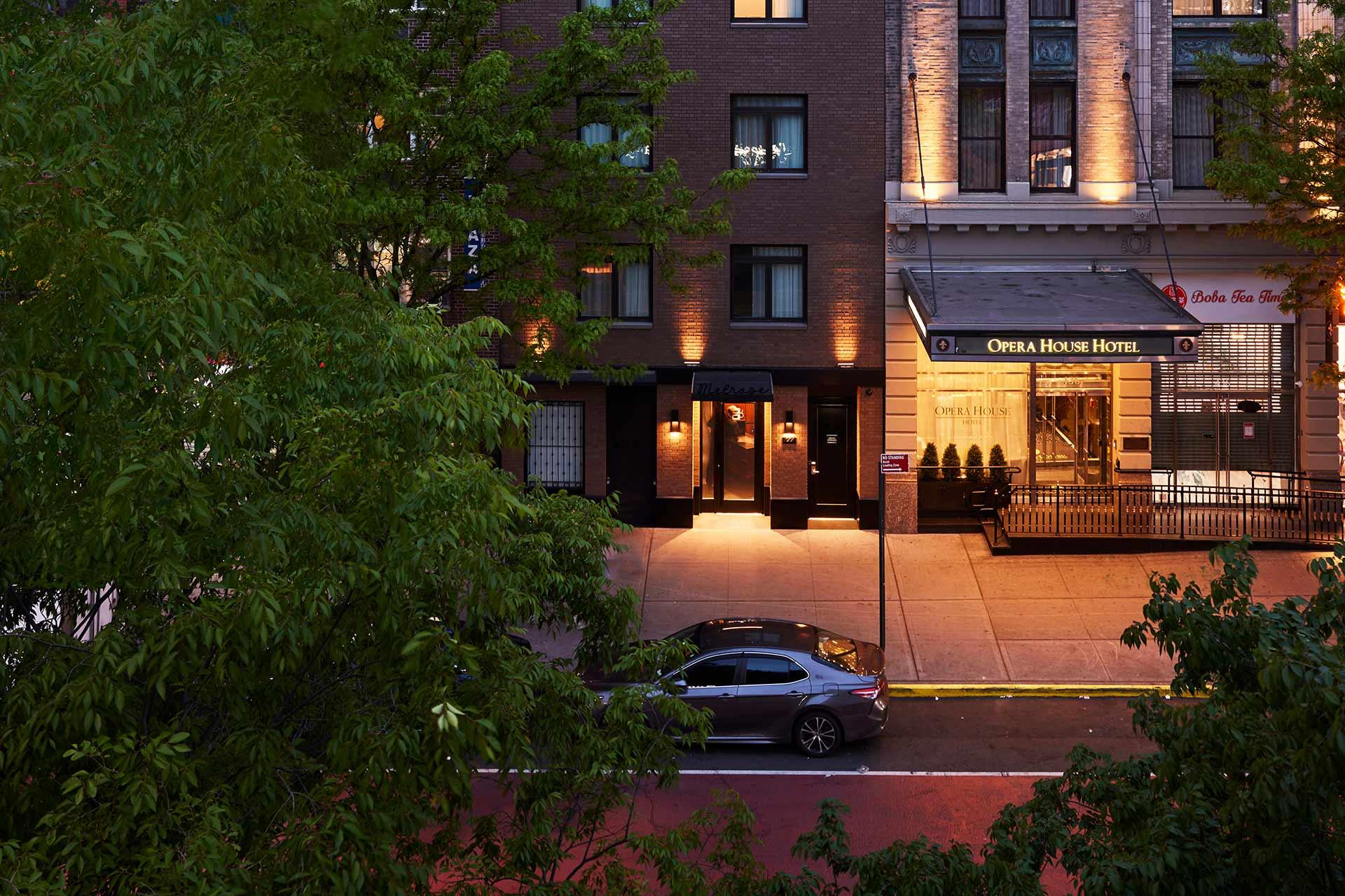 A car parked on a city street in front of the lit entrance to the Opera House Hotel, with trees partially obscuring the view and building facades illuminated at dusk.