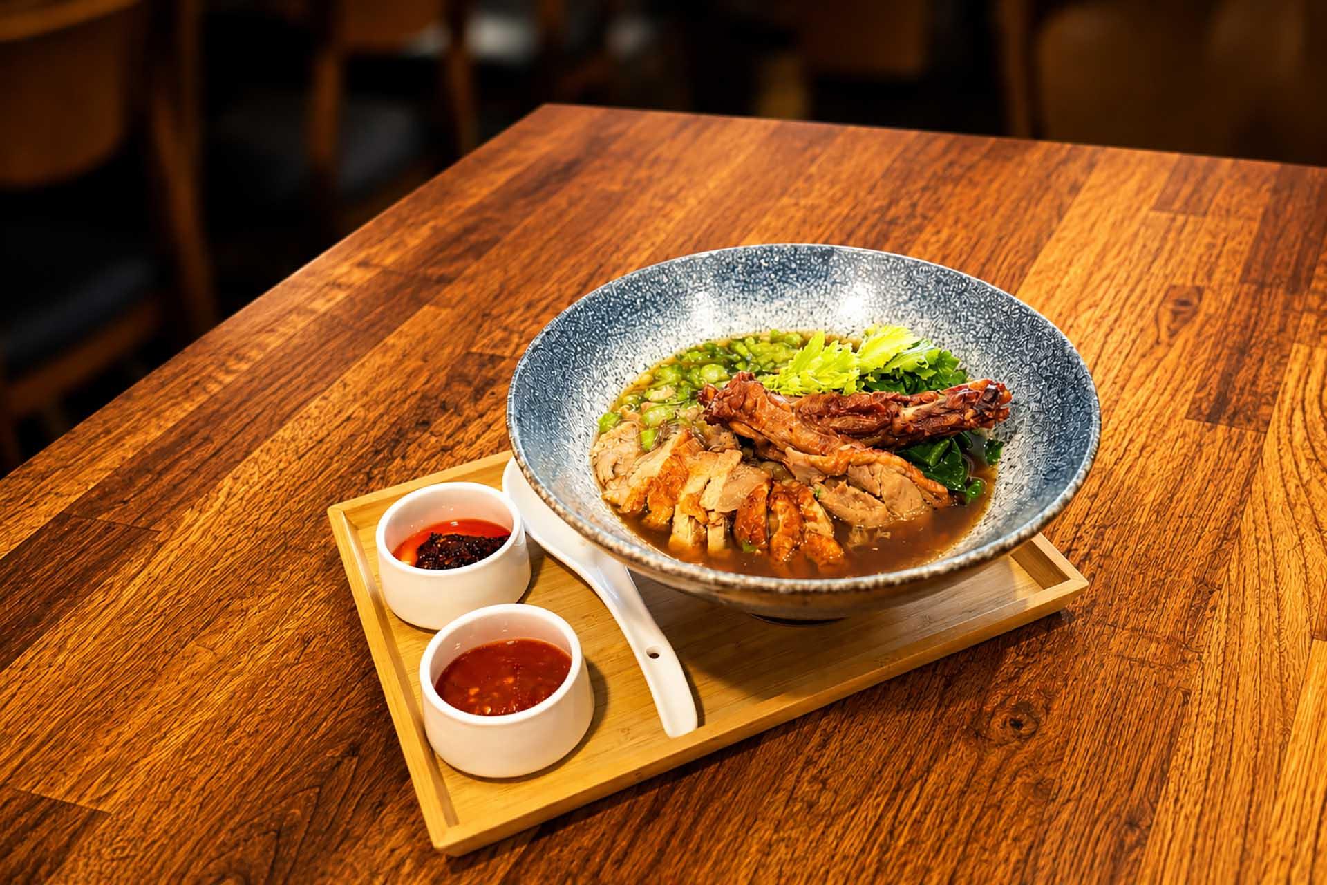 A bowl of noodle soup with sliced meat and greens, served on a wooden tray with two small bowls of red sauces and a white spoon, set on a wooden table.