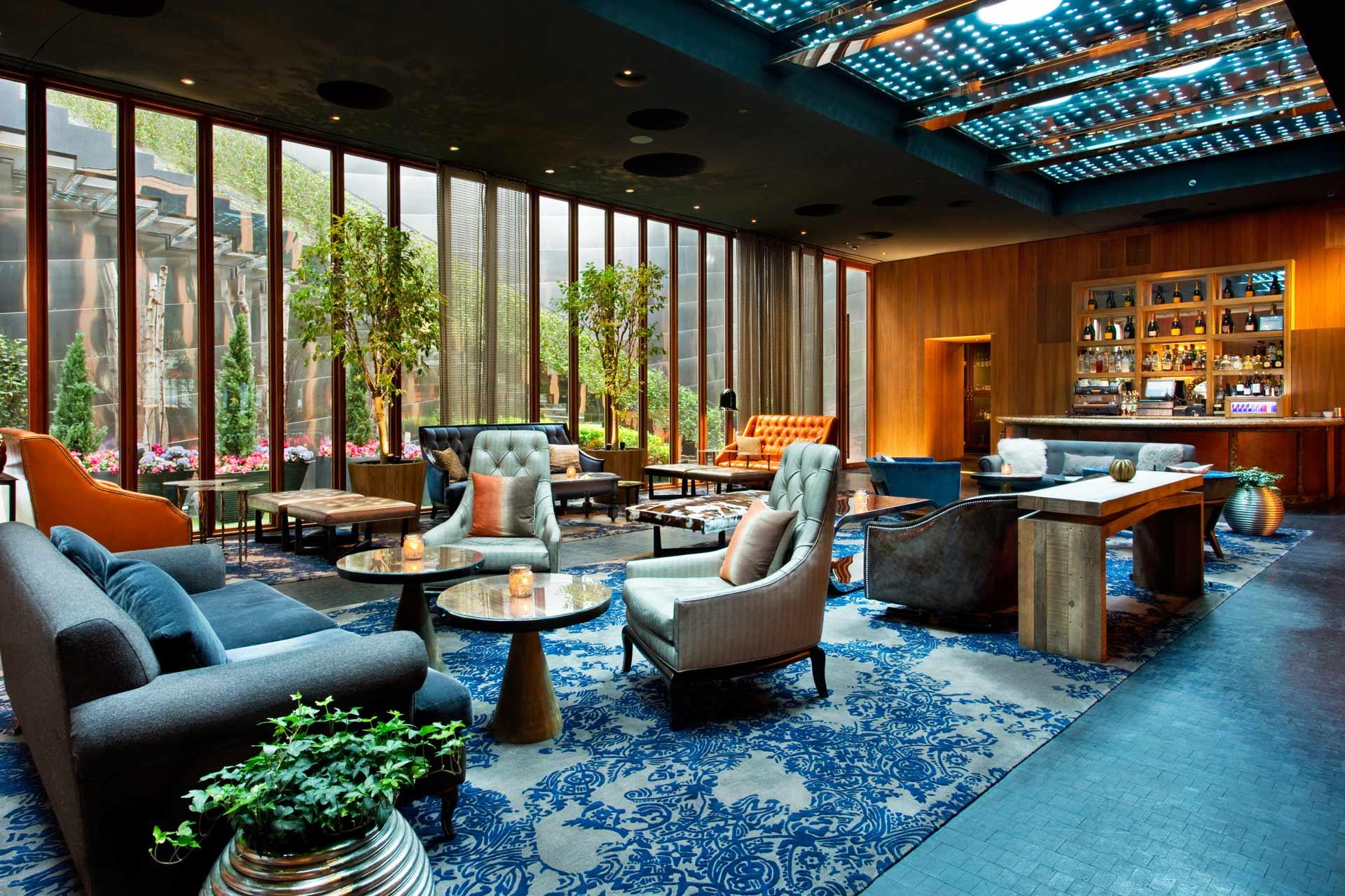 A stylish hotel lounge with modern sofas, armchairs, tables, and a patterned blue carpet. Large windows let in natural light and greenery. There is a bar area in the back and potted plants throughout the space.