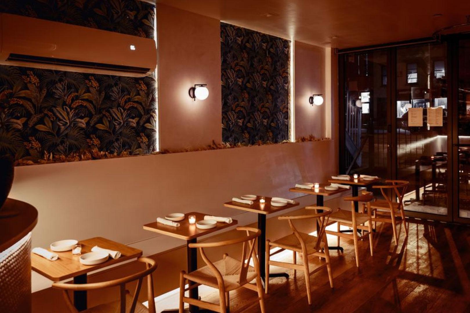 A cozy restaurant interior with wooden tables and chairs, dim lighting, patterned wallpaper, wall sconces, and a row of place settings. Warm tones and a modern, inviting atmosphere.