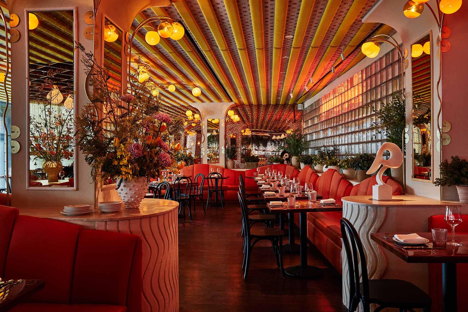 A stylish restaurant with red cushioned seating, dark wood tables, floral arrangements, striped ceiling, warm lighting, and decorative mirrors creating a cozy, vibrant atmosphere. Tables are set and ready for diners.