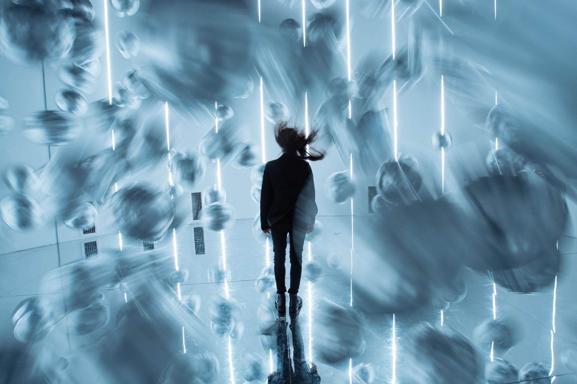 A person stands in a futuristic, glowing room while blurred spherical objects rush past them, creating a dynamic, surreal atmosphere.