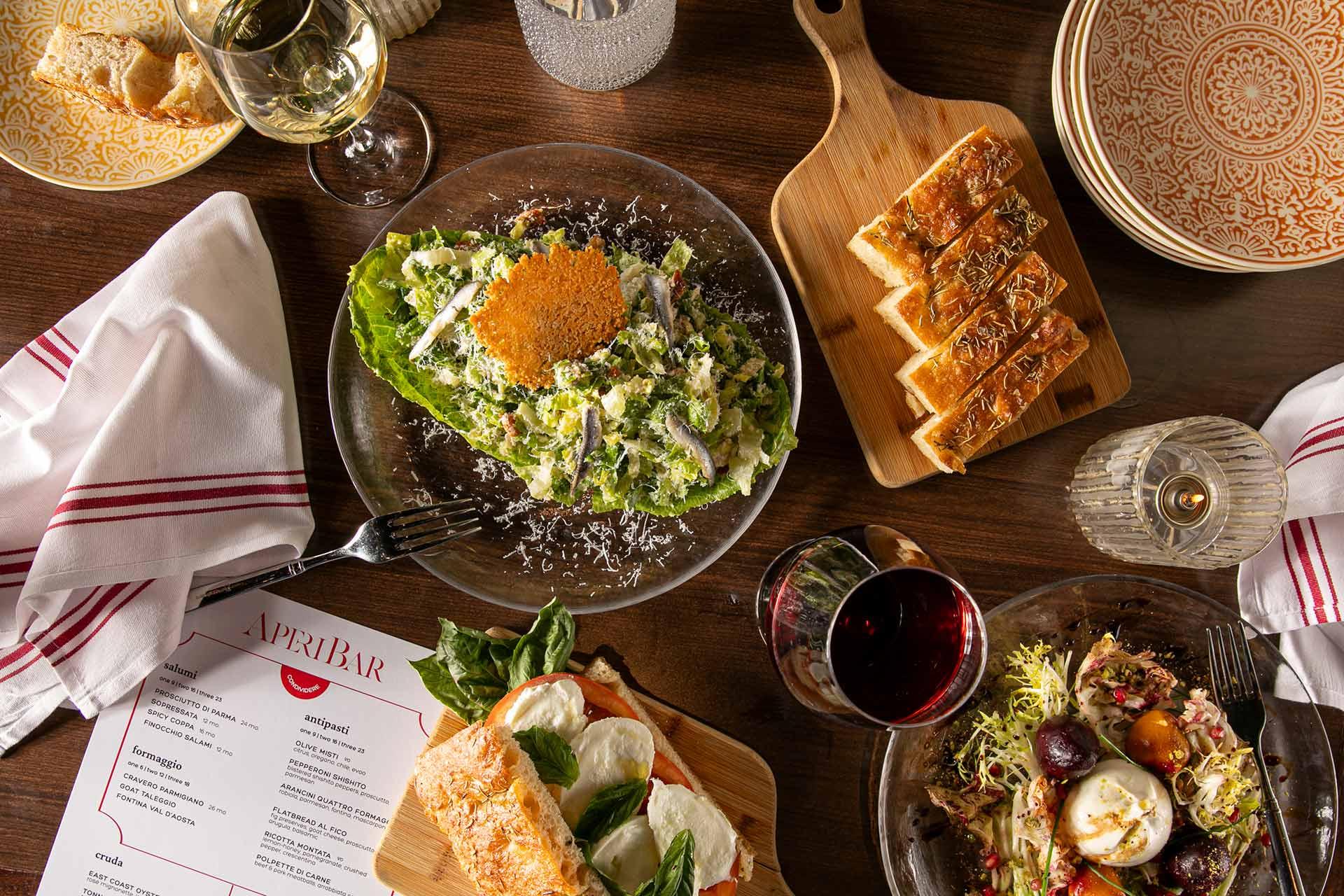 A table set with various dishes including Caesar salad, sliced bread on a wooden board, a Caprese salad, a mixed greens salad, glasses of white and red wine, a menu labeled "APRILBAR," and patterned plates.