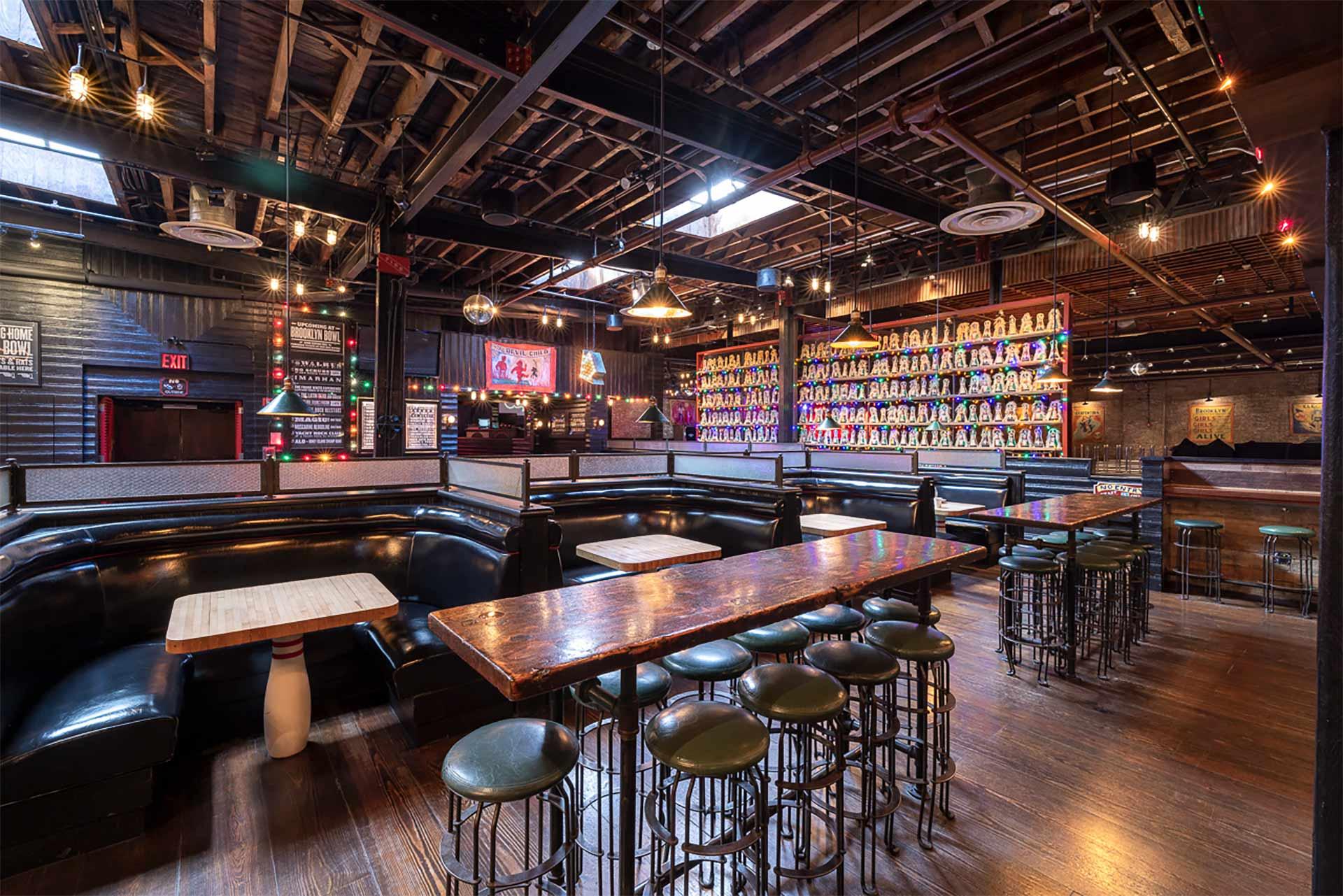 A spacious bar with wooden floors, long tables, black leather booths, and high stools. One wall displays many colorful matryoshka dolls, and string lights hang from the exposed ceiling beams.