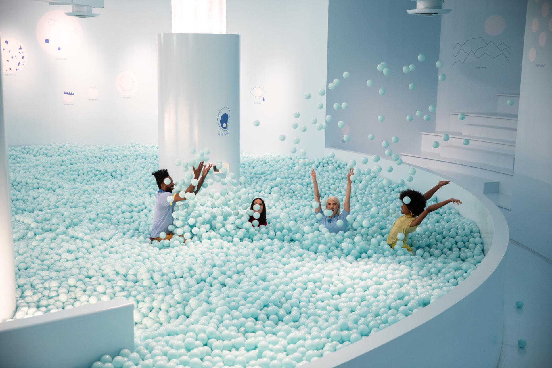 Four children play and throw light blue balls in a large indoor ball pit with white walls and soft lighting, creating a joyful and lively atmosphere.