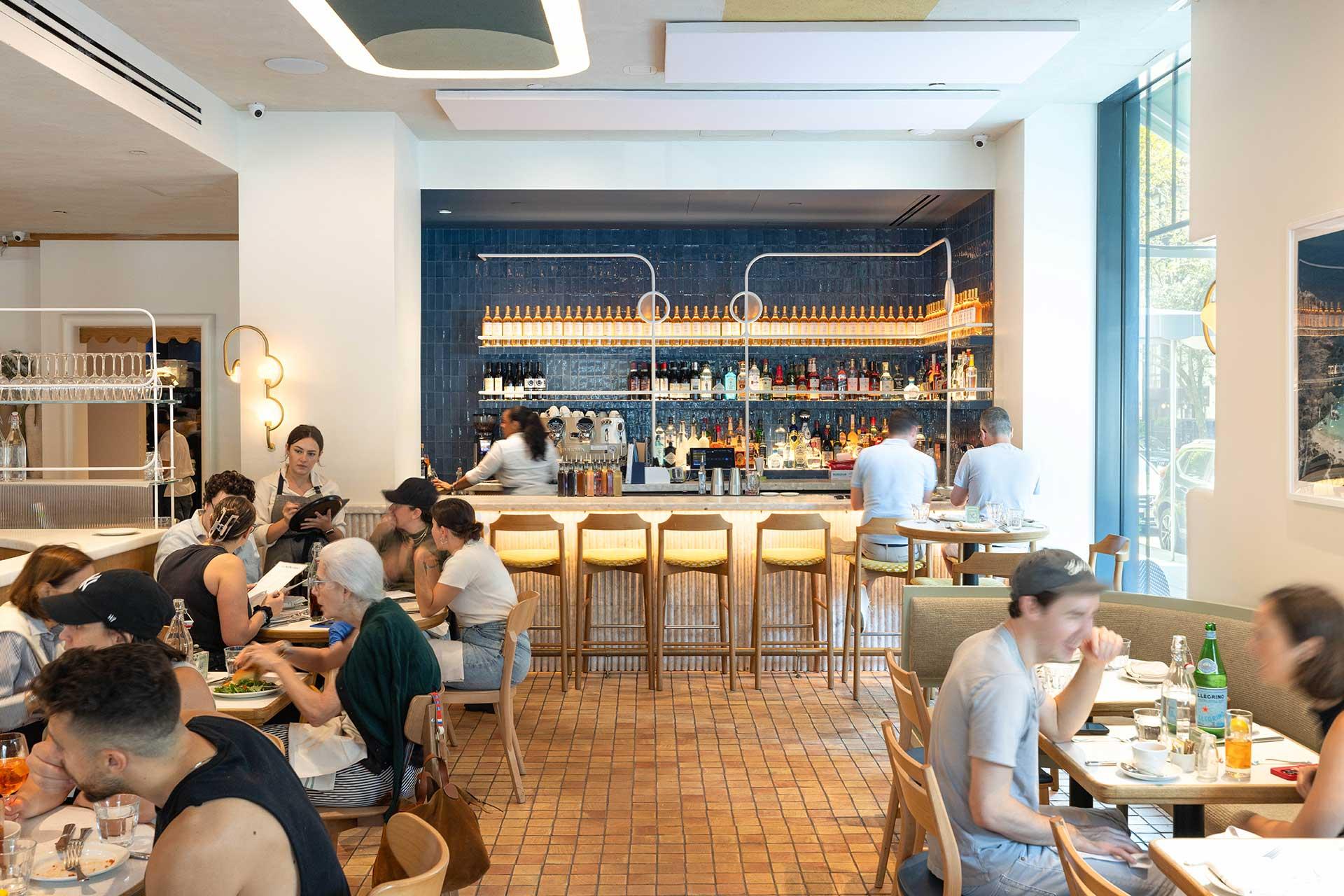 A bright, modern restaurant with people dining at tables and a bar in the background. The space features wooden furniture, a tiled floor, large windows, and shelves with bottles behind the bar.