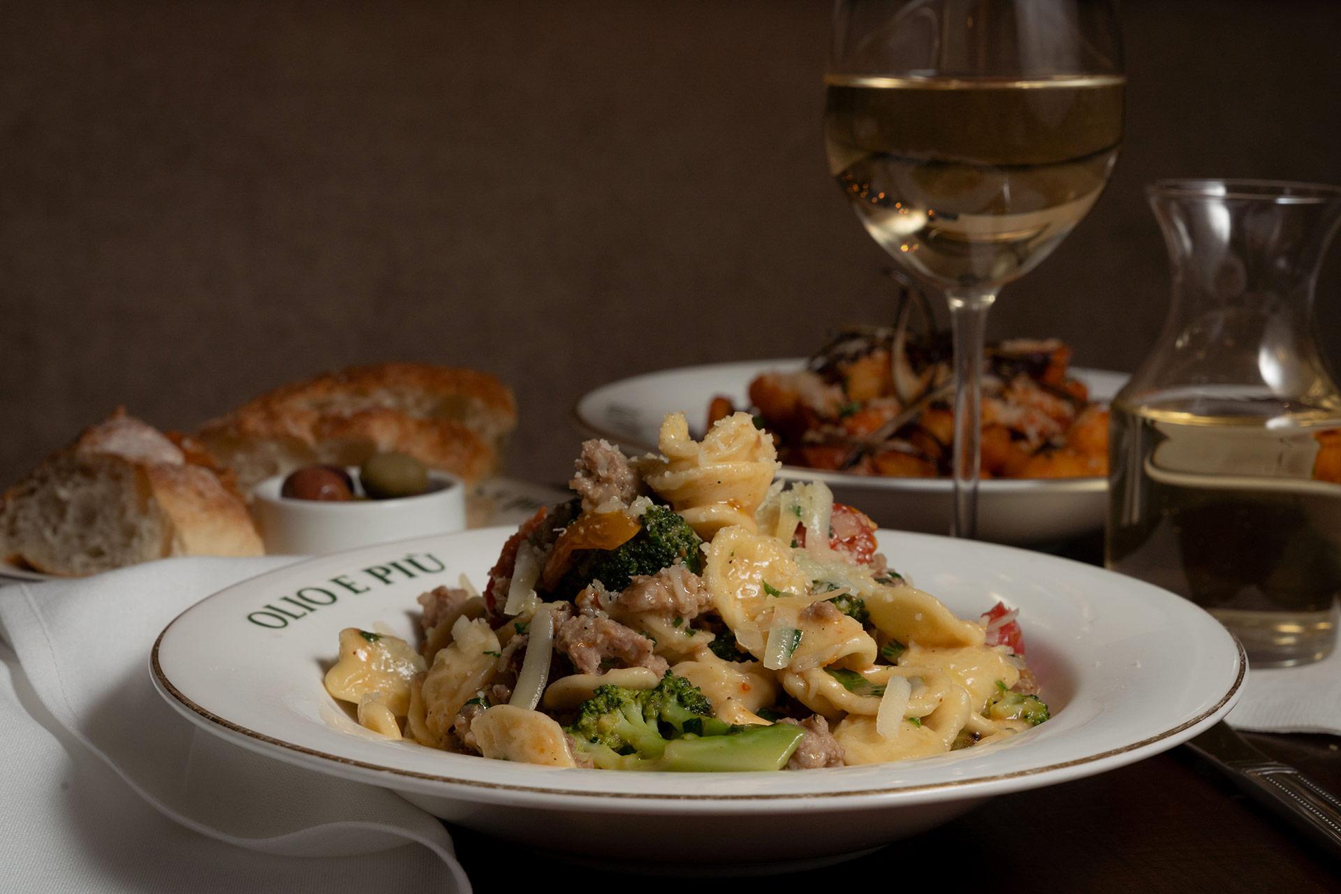 A plate of pasta with sausage, broccoli, and cheese sits on a table next to a glass of white wine, a carafe, a bowl of bread, and a small dish of olives. The plate is labeled "Olio e Più.
