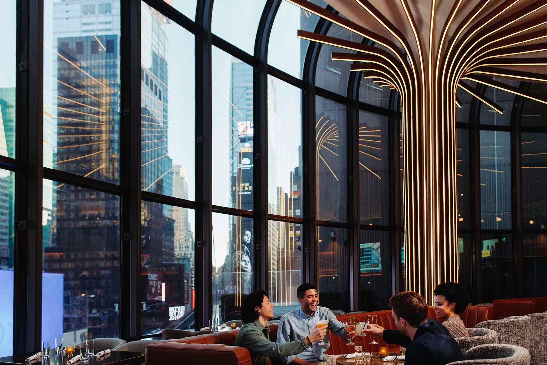 Four people sit around a table in a modern restaurant with tall curved windows, toasting drinks. City skyscrapers and bright lights are visible outside, and stylish lighting decorates the ceiling.