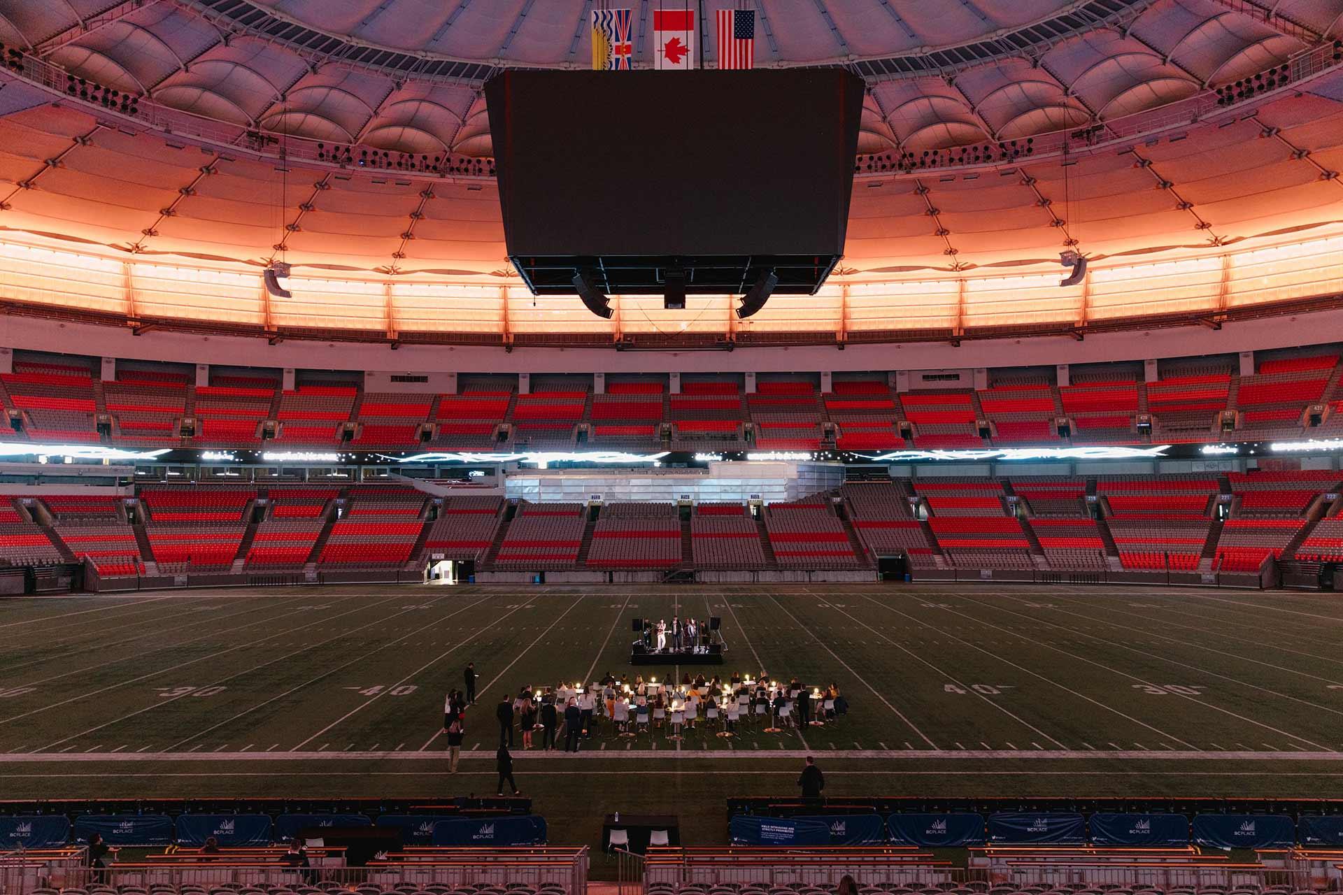 A small orchestra performs on a stage at the center of an empty, brightly lit indoor stadium with red and gray seats and a large screen hanging above the field.