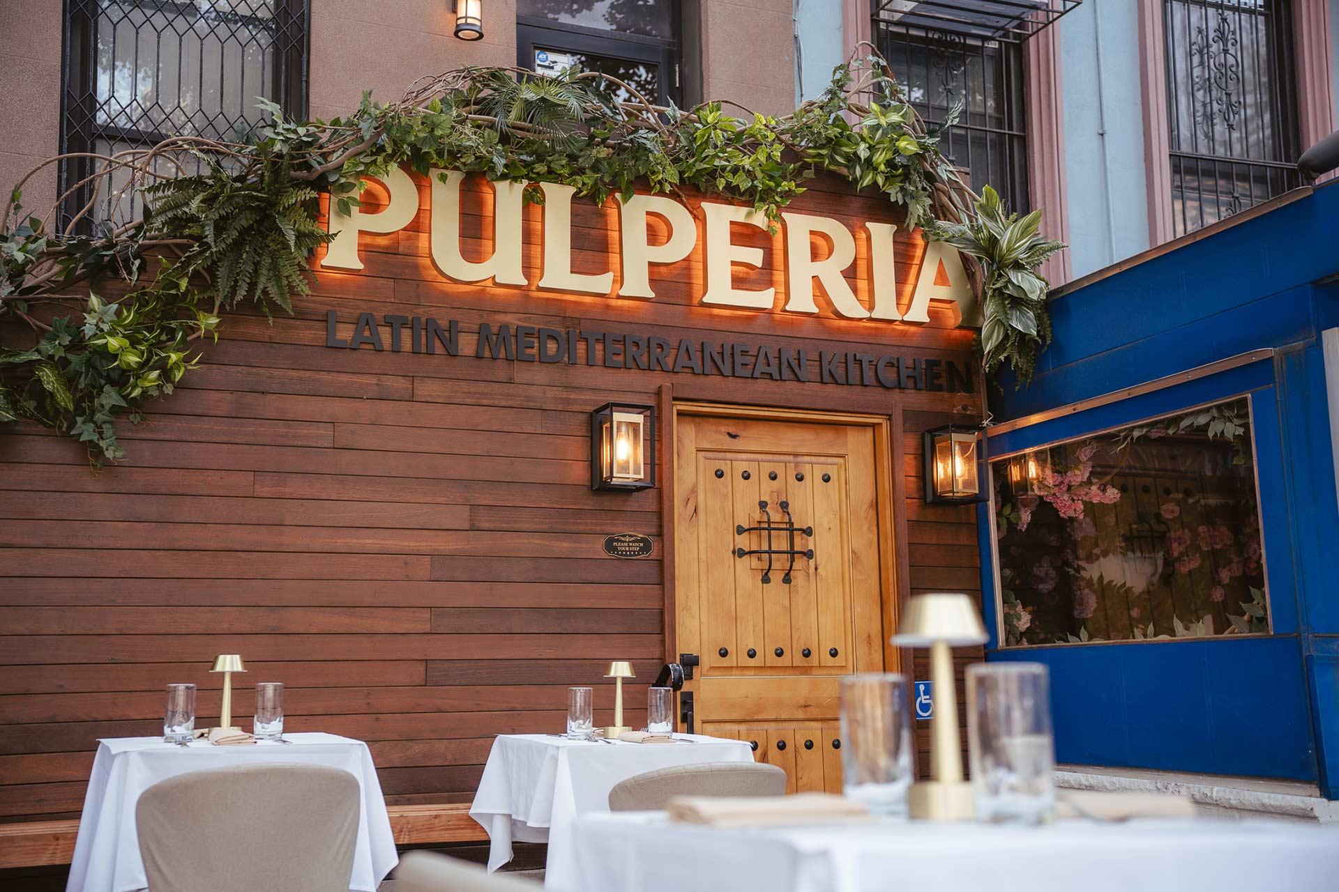 Outdoor seating area of a restaurant with white tablecloth-covered tables in front of a wooden facade that has a sign reading "Pulperia Latin Mediterranean Kitchen" above a wooden door, decorated with greenery.