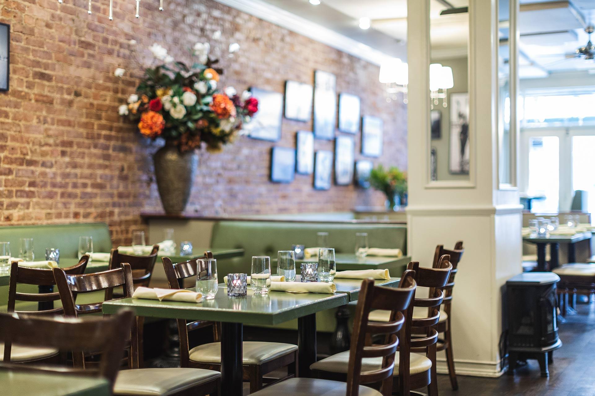 A cozy restaurant with green tables, wooden chairs, and place settings. A large flower arrangement sits against a brick wall decorated with framed photos and artwork. Soft lighting creates a warm, inviting atmosphere.