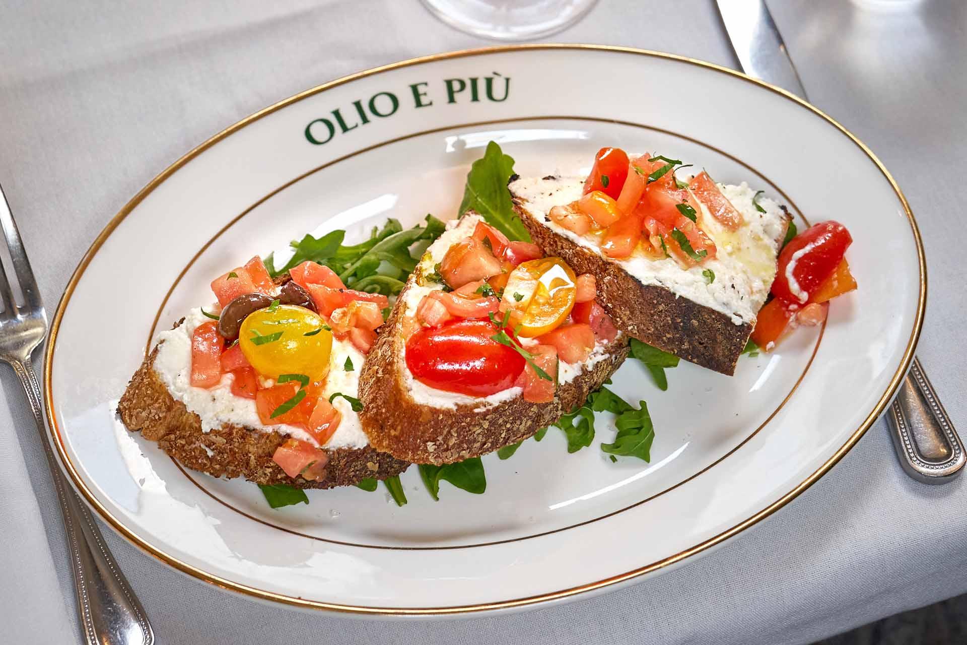 Three slices of bread topped with ricotta cheese, chopped tomatoes, and herbs sit on a bed of arugula on a white plate labeled “OLIO E PIÙ.” Silverware and a white napkin are visible beside the plate.