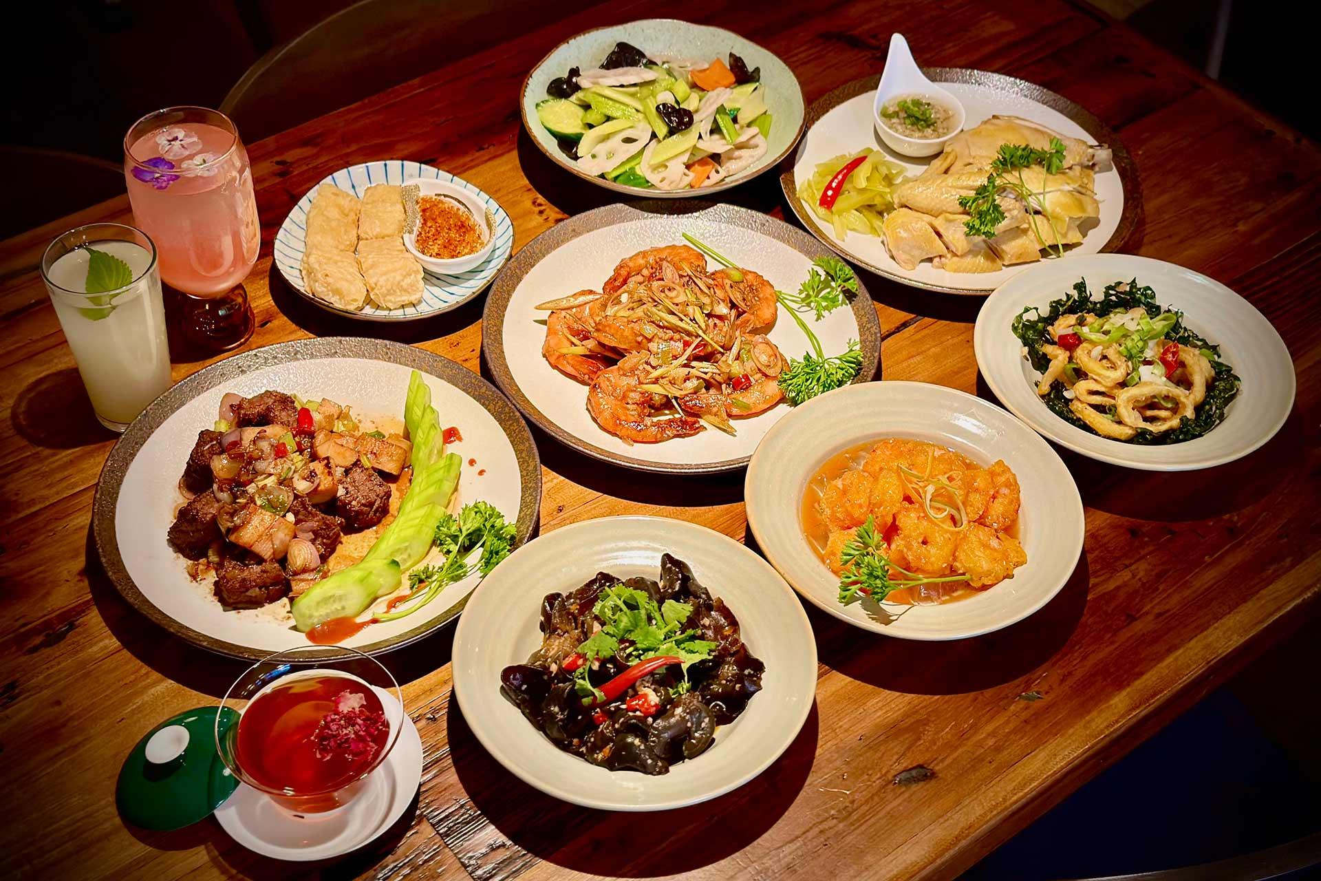 A wooden table filled with various Asian dishes, including shrimp, pork, vegetables, tofu, tea, and colorful drinks, all neatly arranged and garnished with herbs.