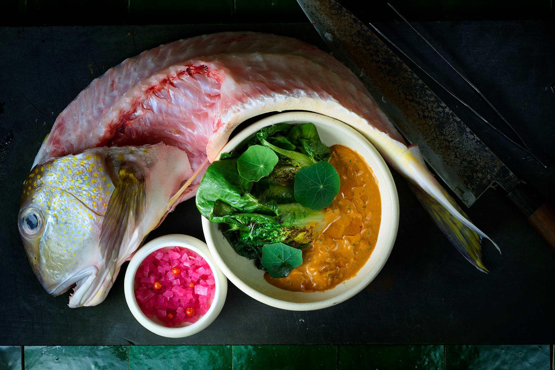 A whole raw fish is curled around a bowl of yellow curry with leafy greens and edible flowers, beside a small bowl of diced pink pickles. A large chef’s knife lies above the arrangement on a dark surface.