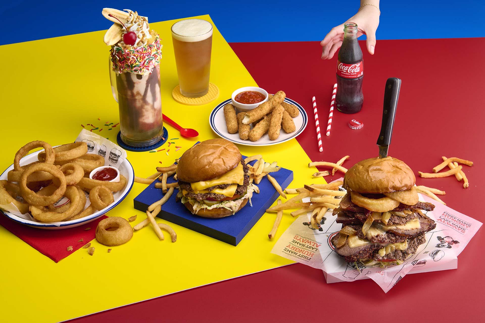 A colorful table with onion rings, fries, cheeseburgers, mozzarella sticks, a milkshake with sprinkles, a beer, and a hand reaching for a glass bottle of Coca-Cola. The scene is playful and vibrant.