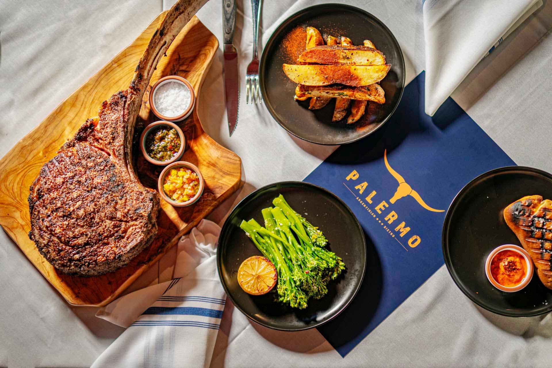 Tomahawk steak on a wooden board with small bowls of seasoning. Plates of potato wedges and broccolini arranged nearby on a table with a blue restaurant menu labeled "Palermo." A knife, fork, and napkin complete the setting.