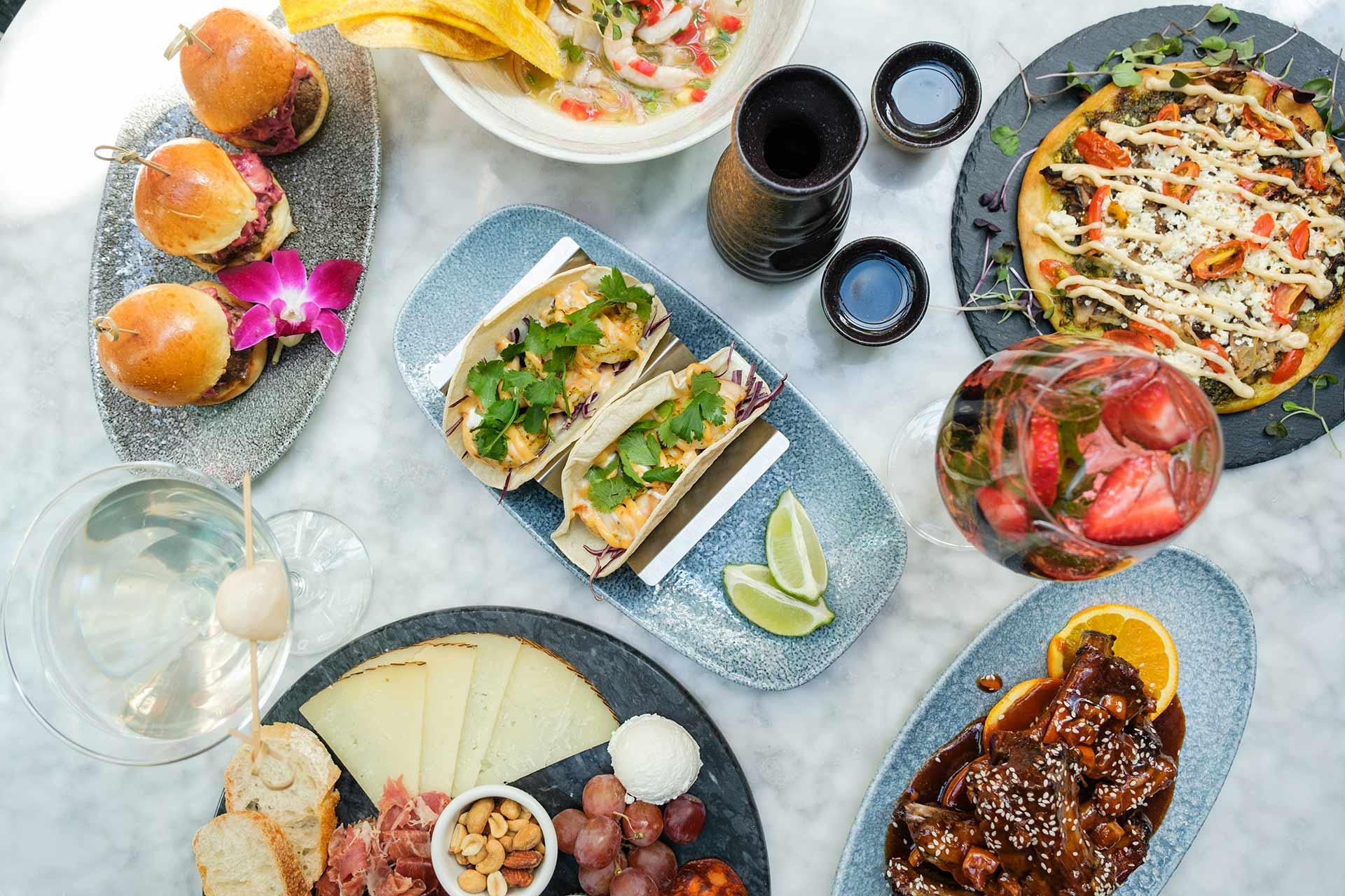 A marble table set with assorted dishes including sliders, tacos, cheese and charcuterie, flatbread, saucy ribs, ceviche, cocktail, and a glass of sangria garnished with fresh fruit and herbs.