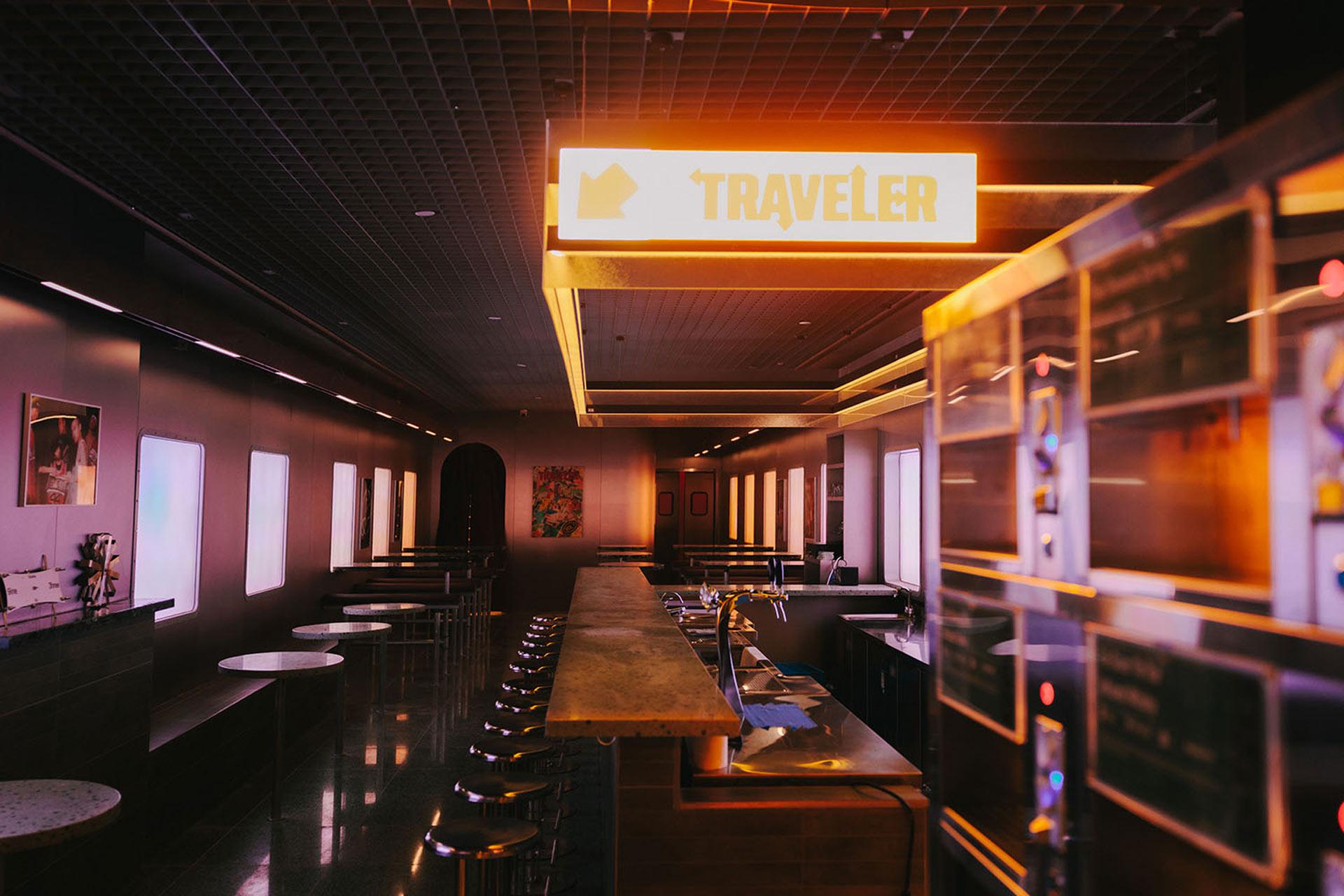A modern, dimly lit café or lounge with marble tables, bar seating, and a glowing yellow "TRAVELER" sign hanging from the ceiling. The space has a calm, inviting atmosphere.