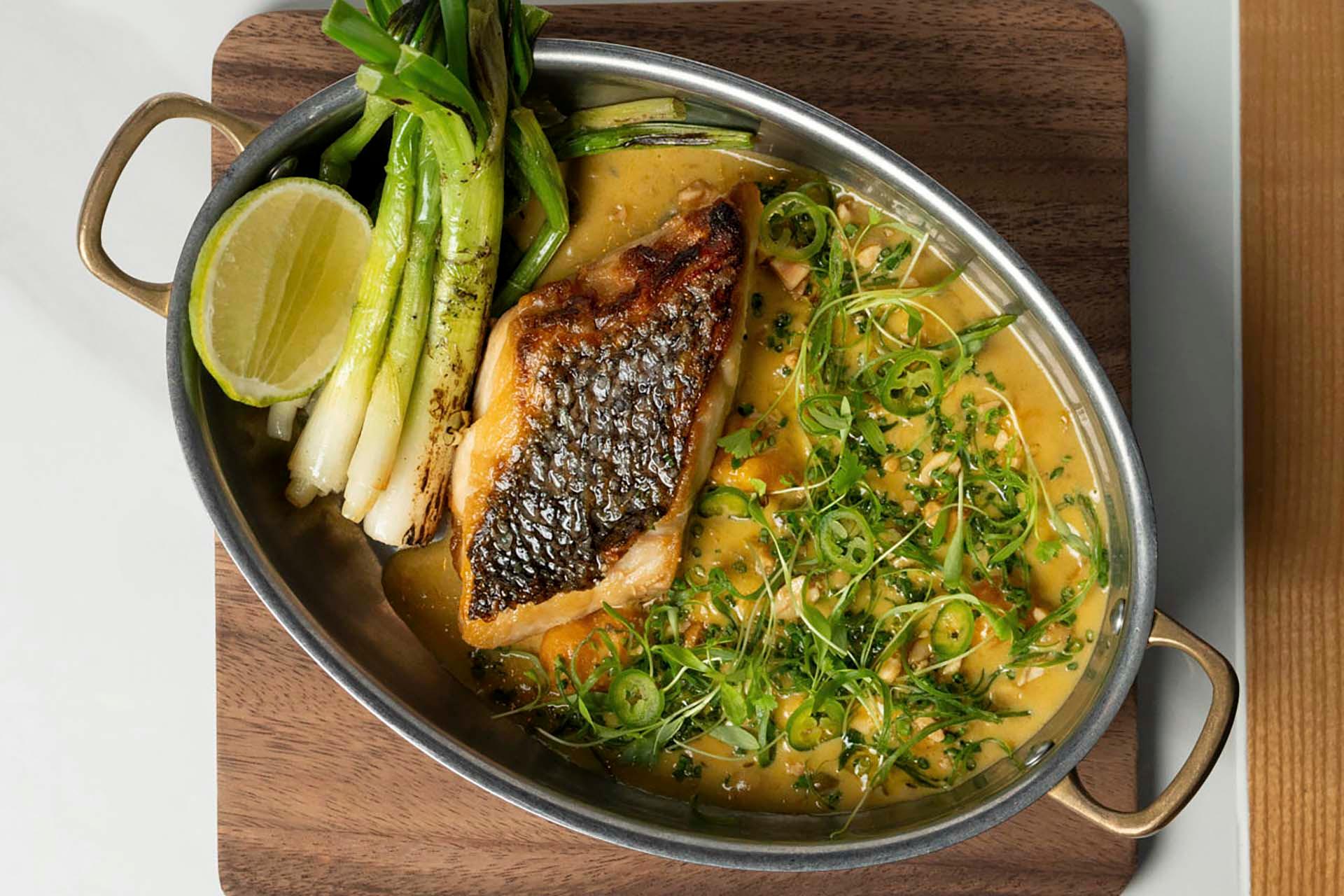 An oval dish with a seared fish fillet on creamy sauce, topped with sliced green chili and herbs. Garnished with grilled green onions and a lime wedge, set on a wooden board.