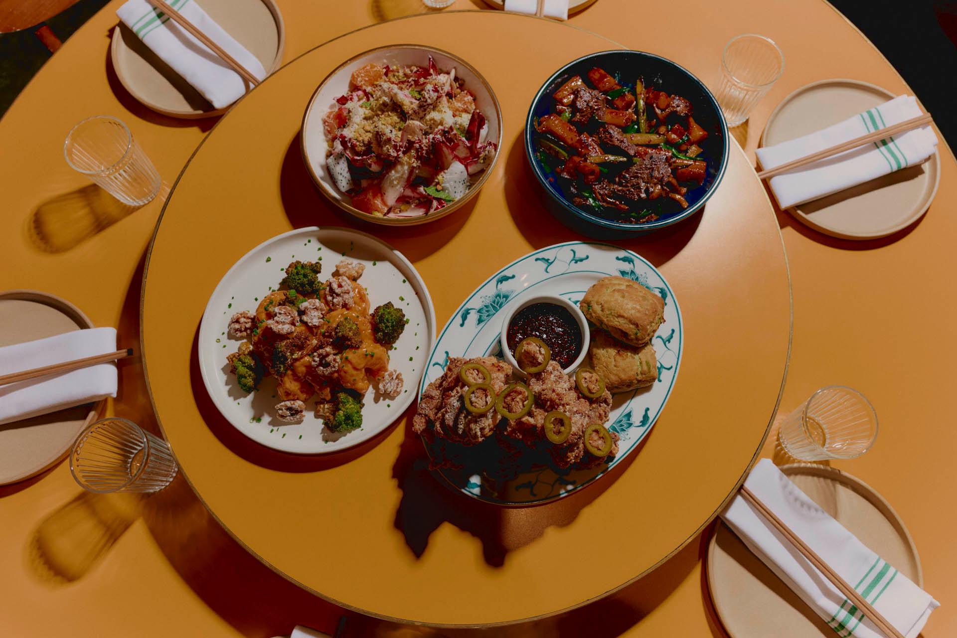 Four plates of assorted food including salad, roasted vegetables, dumplings, and biscuits with a dipping sauce are arranged on a round mustard-yellow table, surrounded by plates, glasses, and napkins.