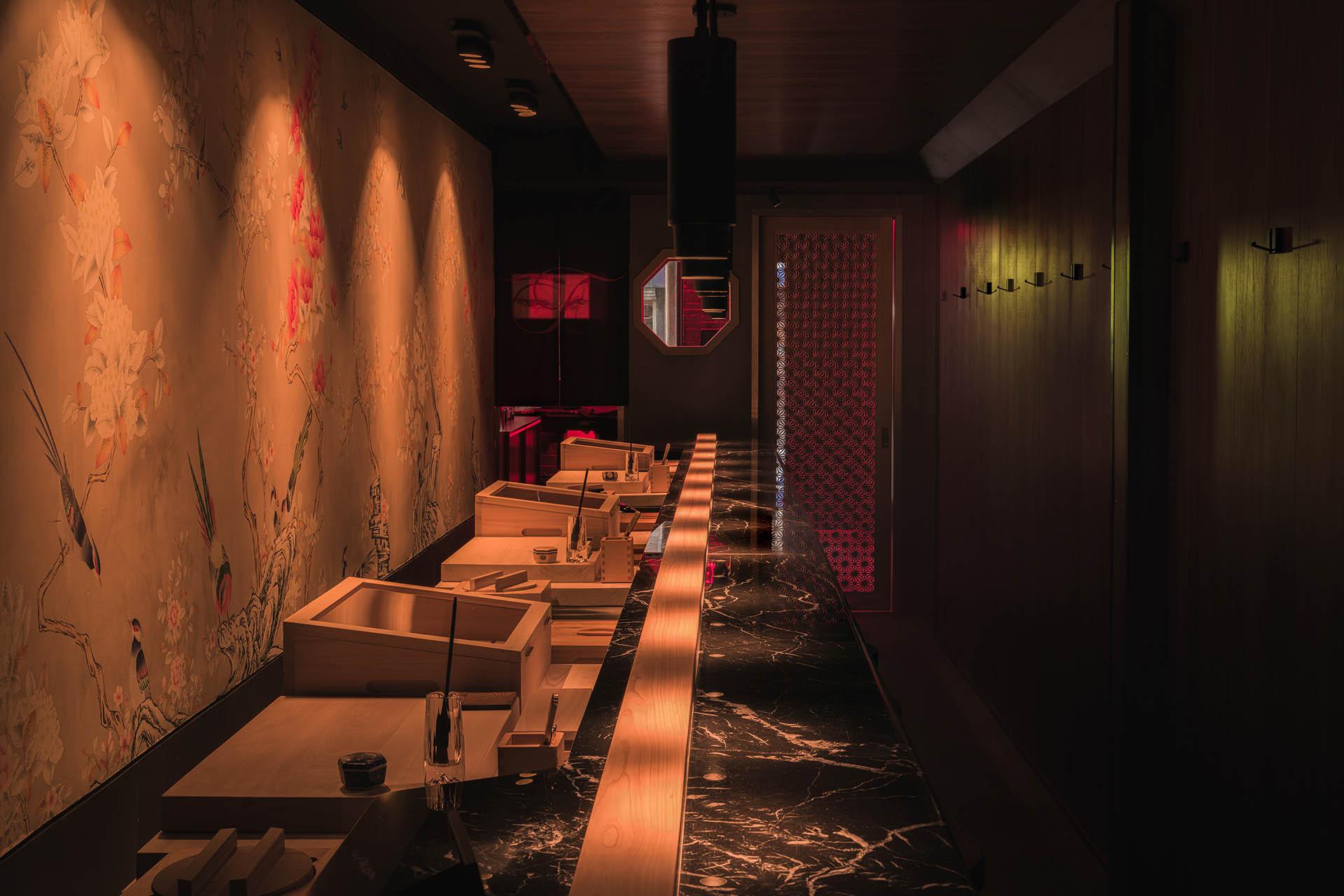 Dimly lit sushi bar with a black marble counter, wooden trays, floral wall art on the left, and a glowing red doorway at the back.