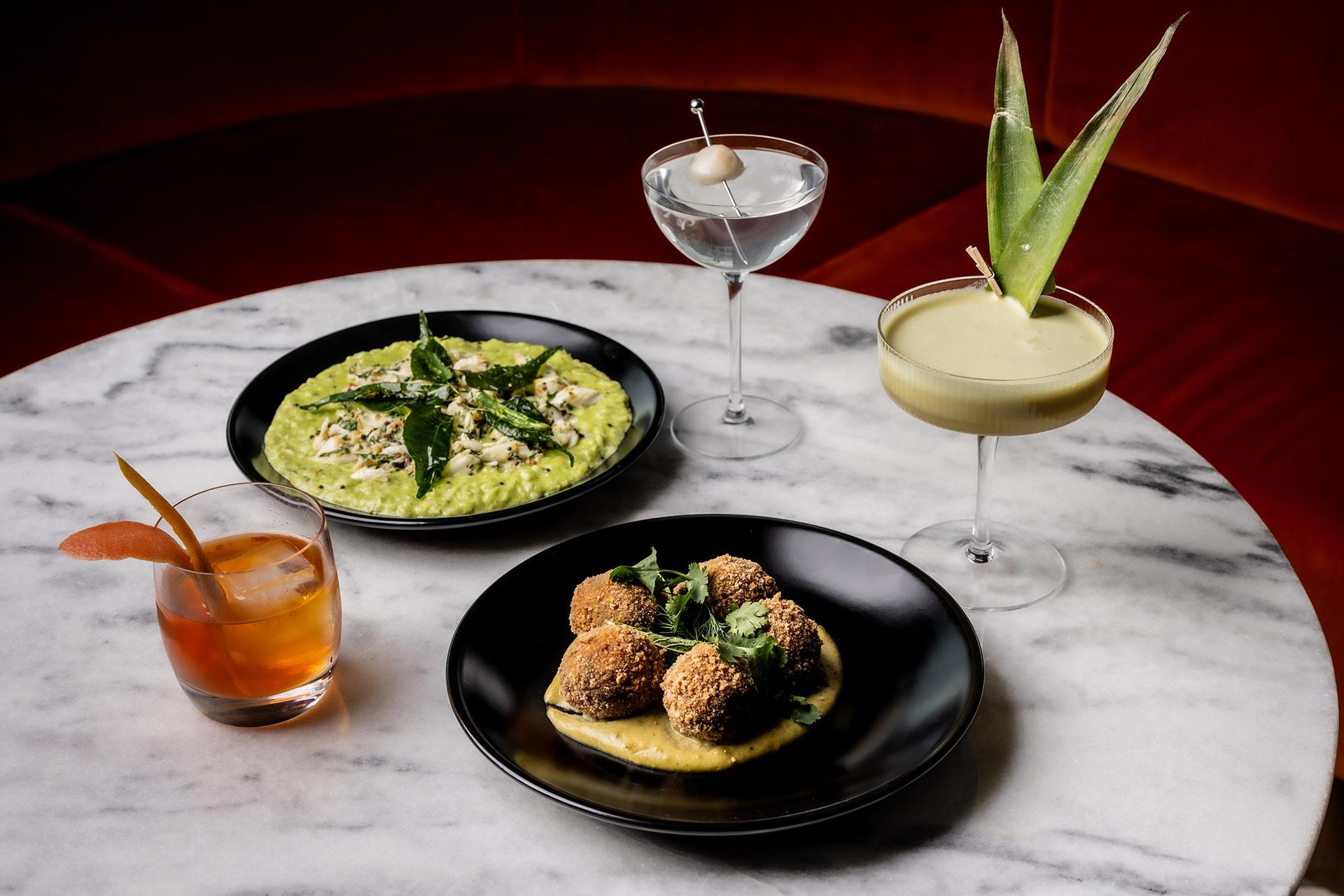 Three cocktails and two plated dishes sit on a round marble table: a Negroni, a martini with an olive, an aloe-garnished cocktail, arancini balls on sauce, and a green risotto topped with herbs and cheese.