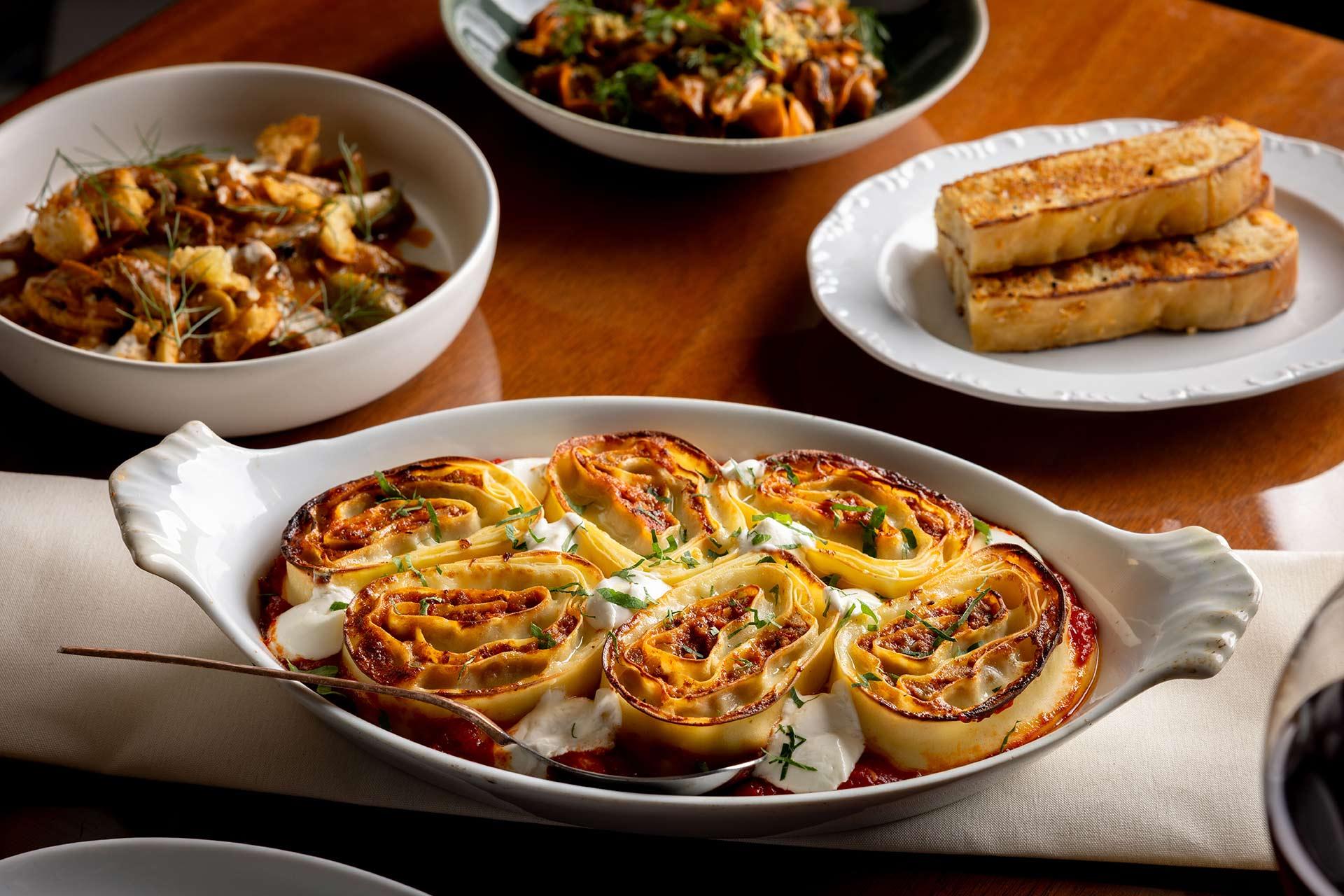 A white dish filled with rolled pasta in tomato sauce and topped with herbs sits on a table, surrounded by grilled bread slices and bowls of other Italian dishes.
