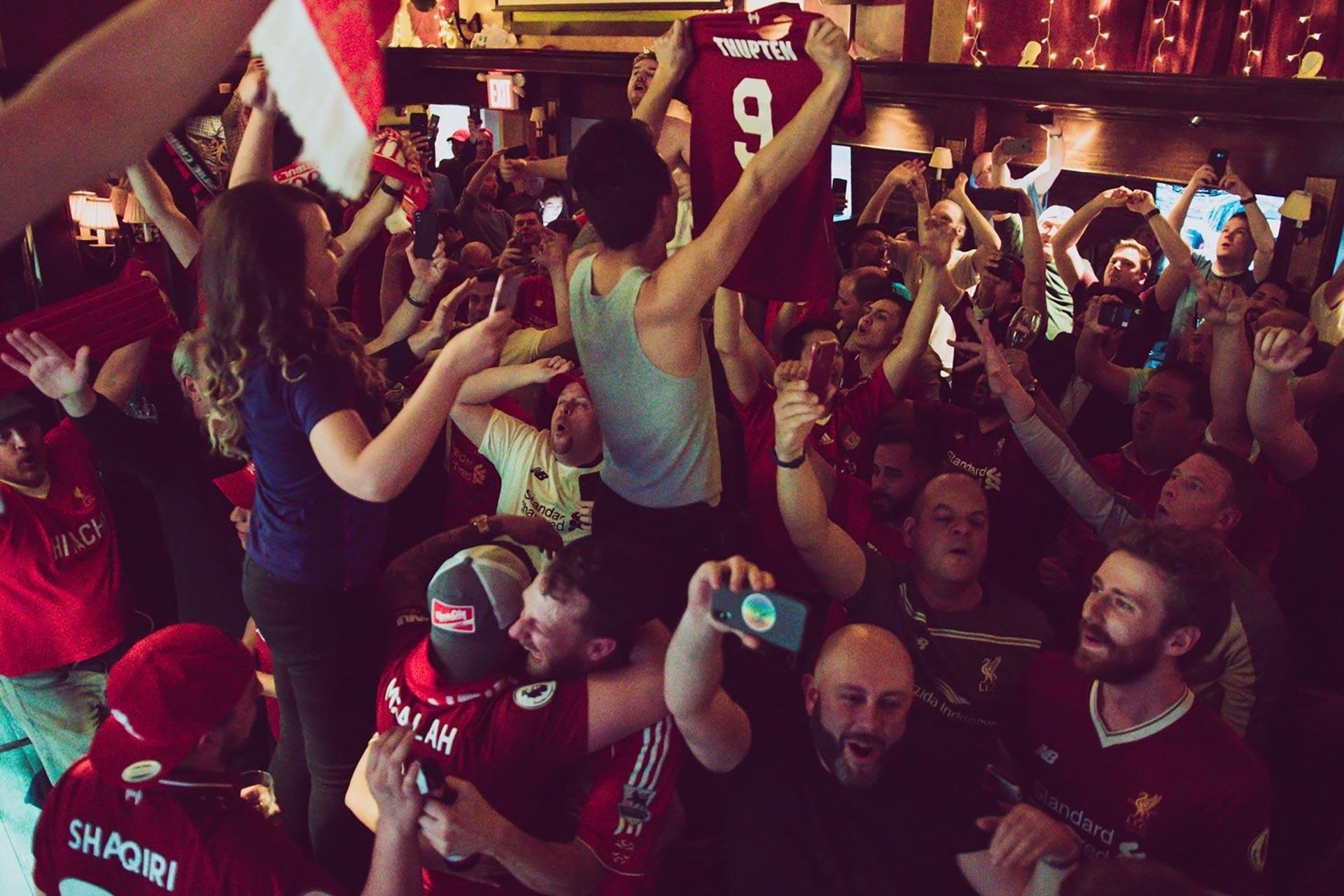 A lively crowd of soccer fans, many wearing red jerseys, cheer and celebrate together in a dimly lit pub. One person holds up a jersey with the name “Firmino” and number 9 as others capture the moment on their phones.