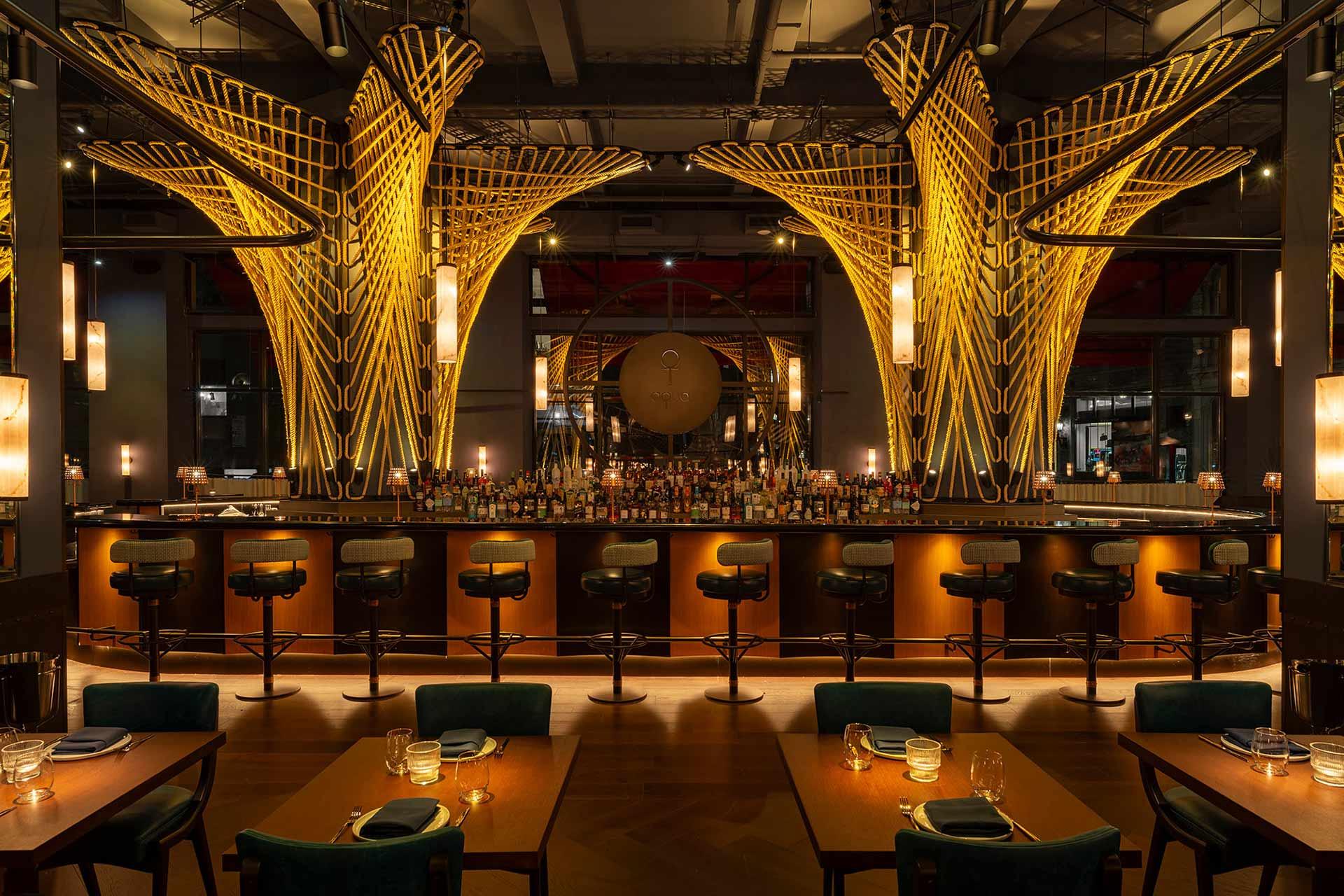 A stylish, modern bar with tall, illuminated golden sculptures behind the counter, high bar stools, warm lighting, and neatly set dining tables in front. Bottles and glasses line the bar, creating an inviting atmosphere.